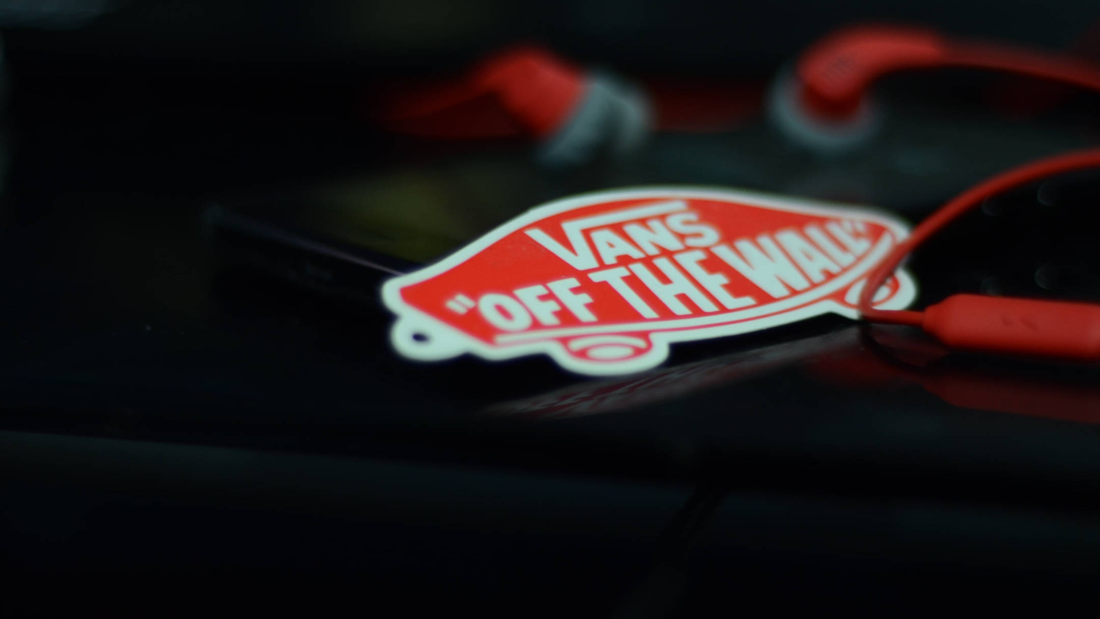Vans Logo 桌布 壁紙 — 一個紅色Vans Off The Wall貼紙放在深色表面上，旁邊有紅色耳機，淺景深。