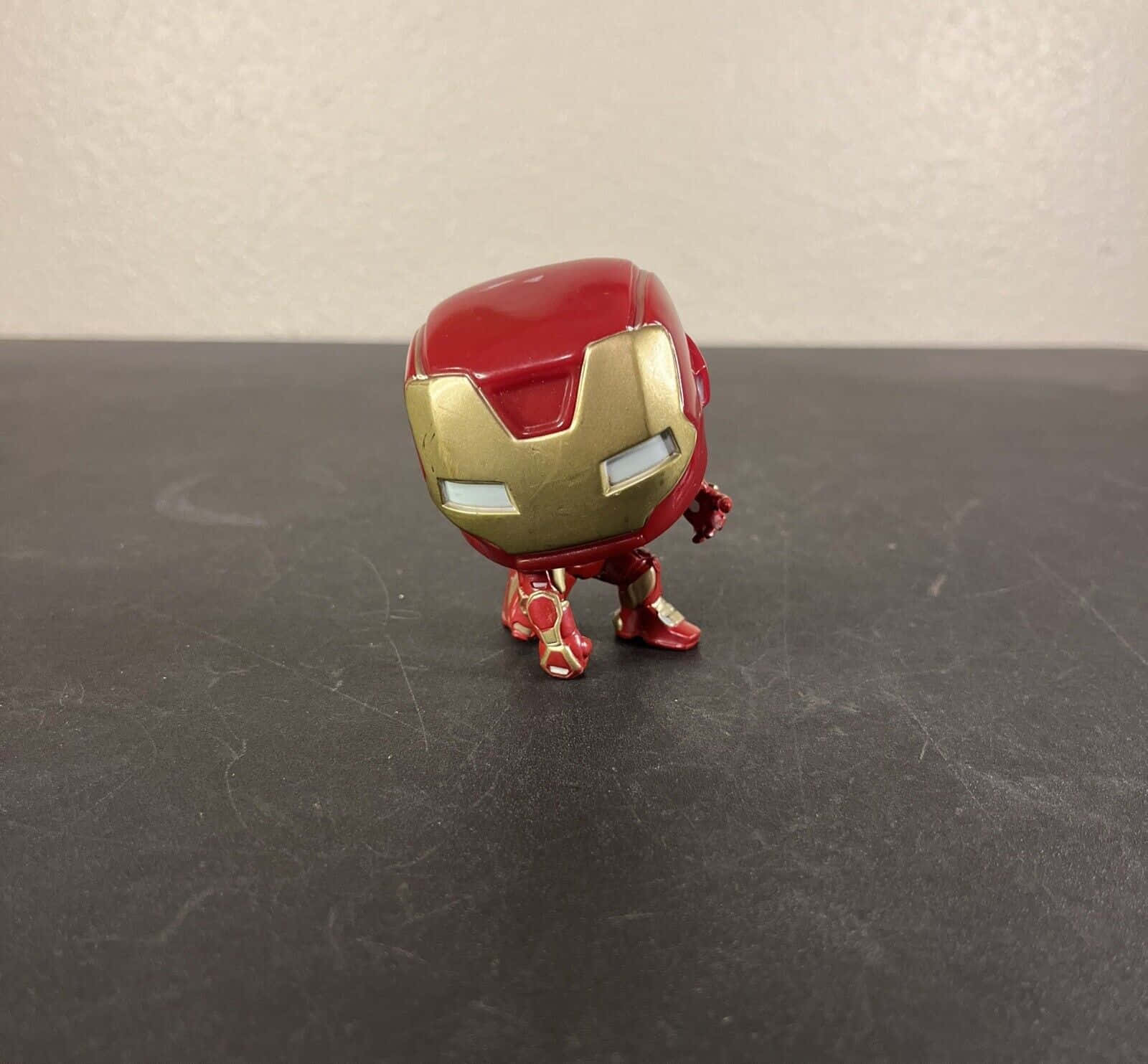 Iron Man Bobbleheads 桌布 壁紙 — 一個紅色和金色的鋼鐵人 Funko Pop 人偶，站立在一個有刮痕的深灰色表面上，背景是純色的米白色牆壁。
