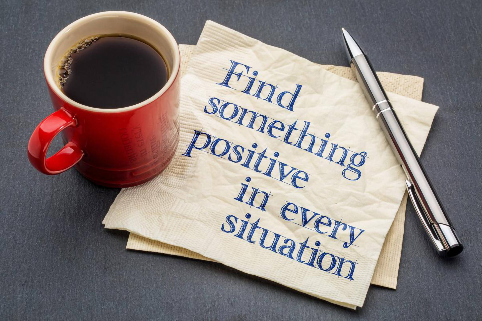Positive Quotes 桌布 壁紙 — 一個紅色咖啡杯放在深灰色表面上，旁邊有一張帶有藍色勵志文字的餐巾和一支銀色筆。