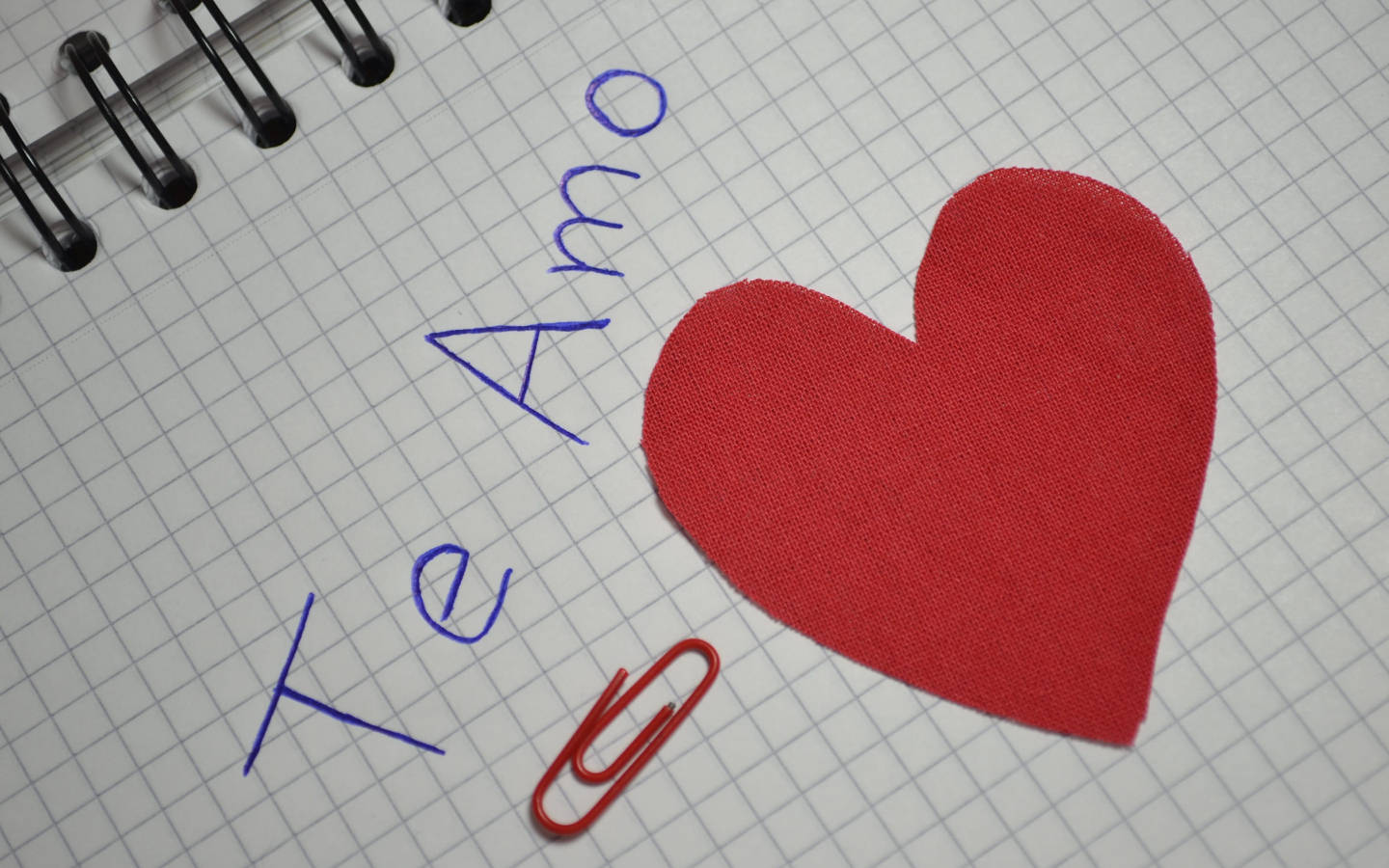 Te Amo 桌布 壁紙 — 一個紅色布料心形和藍色手寫的「Te Amo」放在螺旋筆記本的方格紙上，旁邊有一個紅色迴紋針。