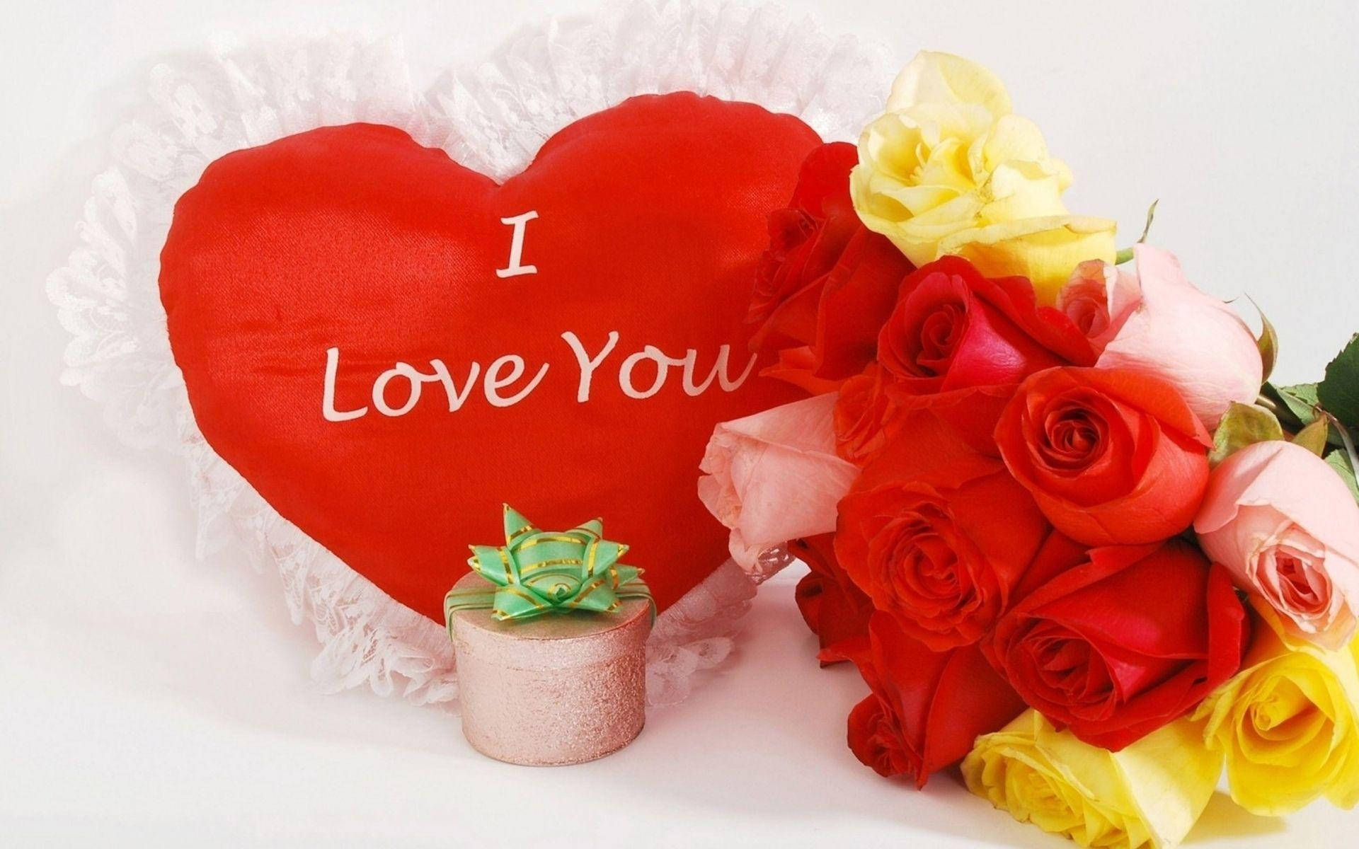 Roses 桌布 壁紙 — 一個紅色心形枕頭，上面有 I Love You 字樣，旁邊是一束鮮豔的紅、粉紅和黃色玫瑰，以及一個小禮物盒。