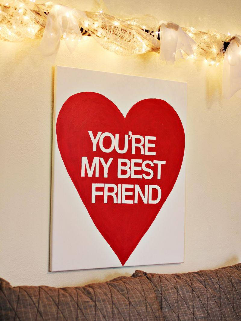 摯友 桌布 壁紙 — 一個紅色心形畫布，上面有白色文字寫著「YOU'RE MY BEST FRIEND」，掛在溫暖的串燈下，下方是棕色沙發。