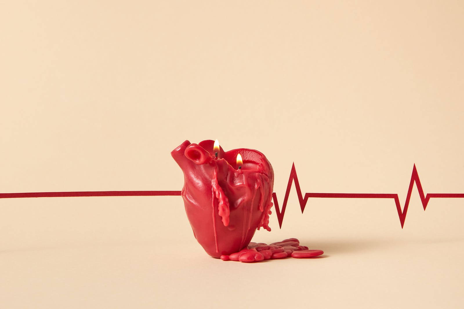 Heartbeat 桌布 壁紙 — 一個紅色心形蠟燭，帶有融化的蠟滴和兩個點燃的燭芯，放在米色背景上的紅色心跳線旁邊。