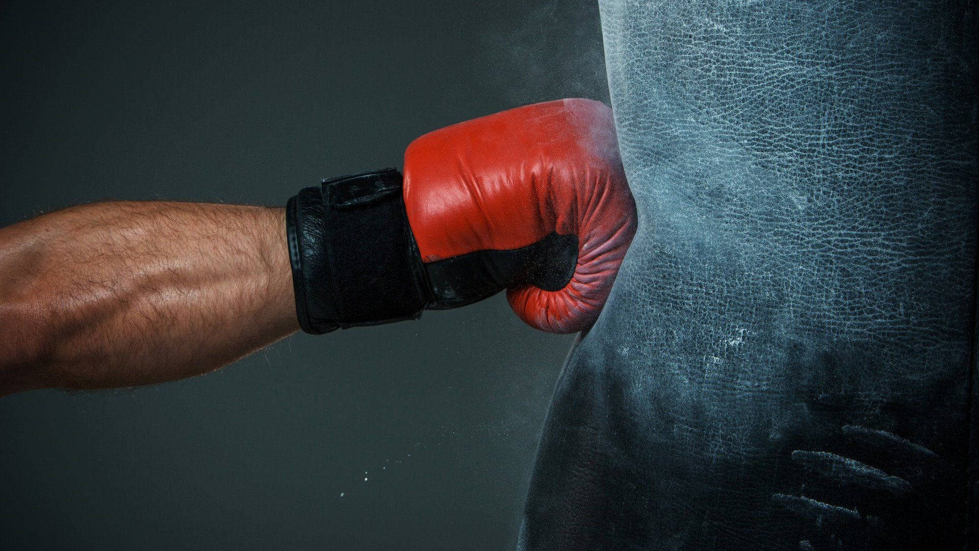 Boxing 桌布 壁紙 — 一個紅色拳擊手套擊打一個磨損的黑色沙包，帶有可見的撞擊灰塵和皮革質感。
