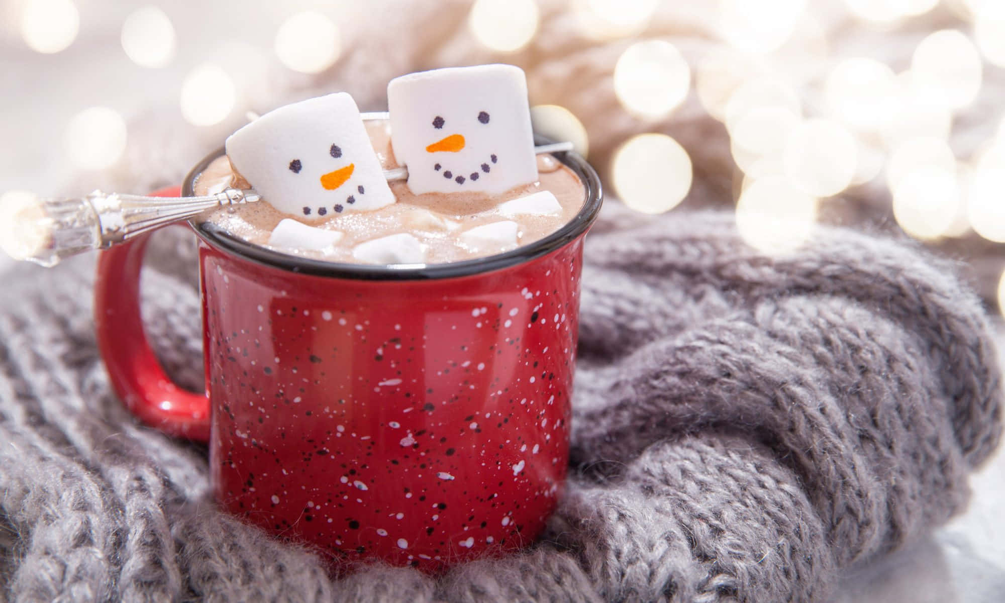 Hot Chocolate 桌布 壁紙 — 一個紅色斑點馬克杯裝滿熱可可和微笑的棉花糖雪人，舒適地放在灰色針織毯上，背景有閃爍的仙女燈。