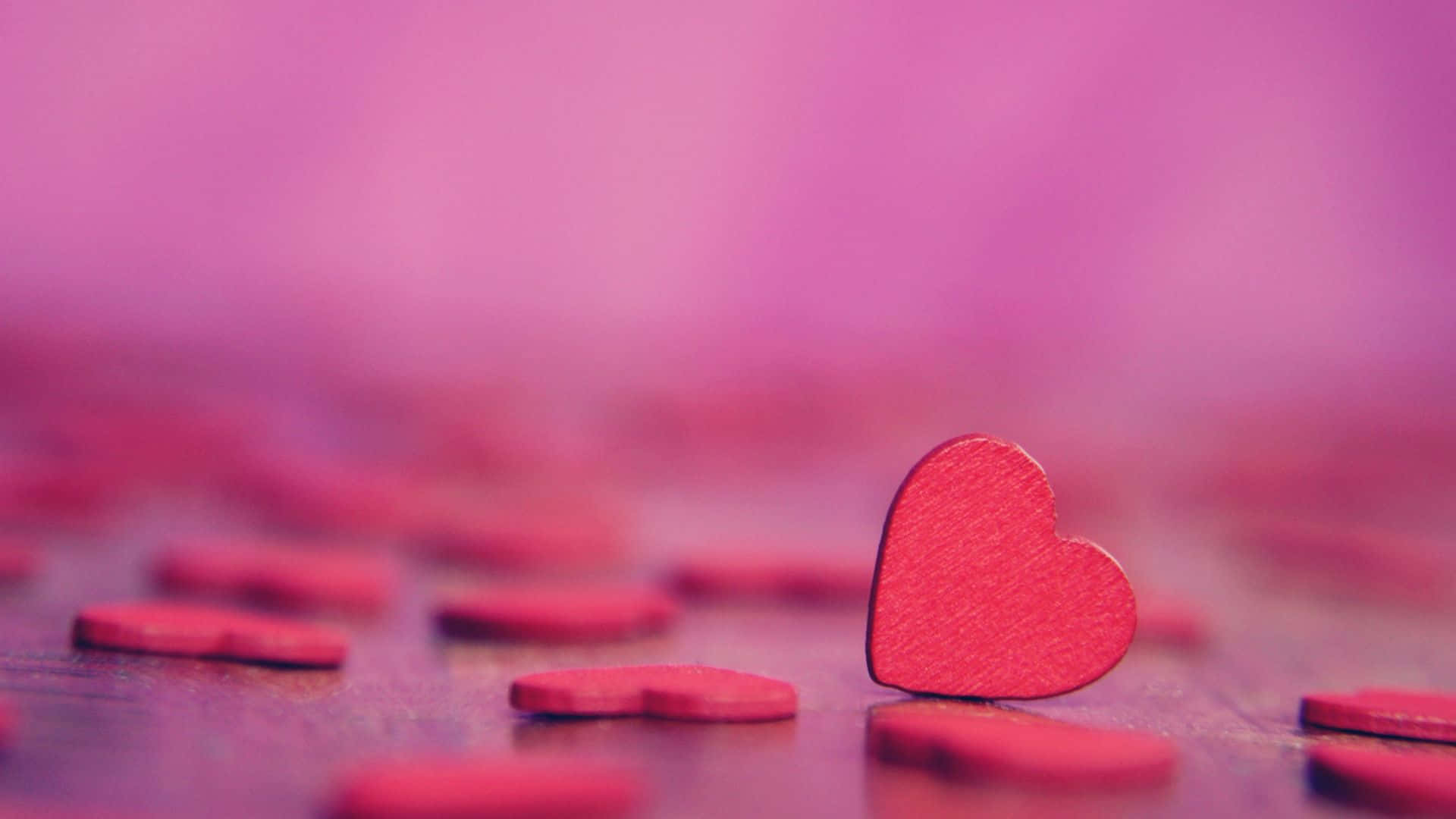 Pink Love 桌布 壁紙 — 一個紅色木心在散落的心形中直立於木質表面上，沐浴在柔和粉紅光中。