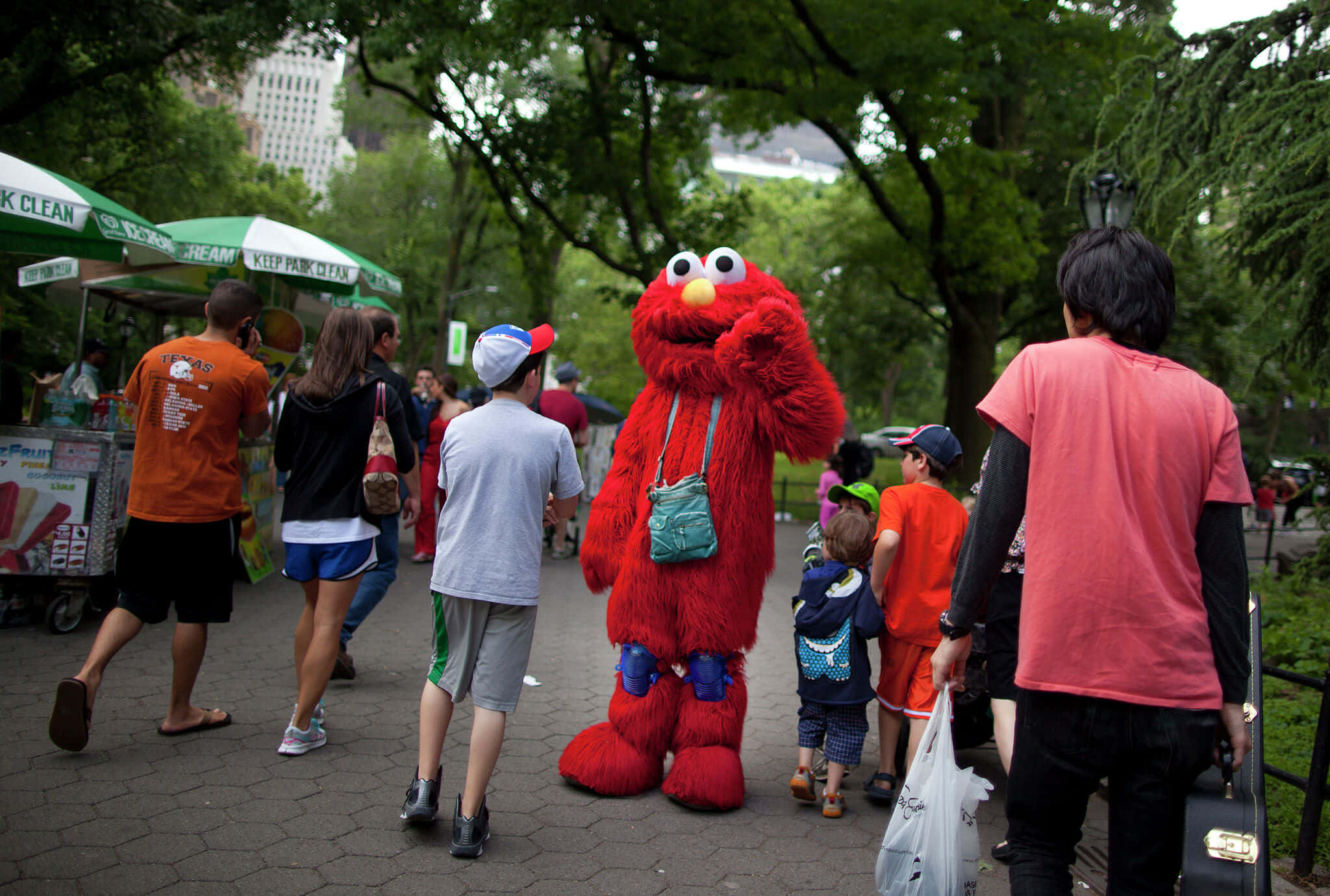 Elmo 桌布 壁紙 — 一個紅色毛茸茸的 Elmo 吉祥物站在繁忙的城市公園裡，人們走過，孩子們圍攏過來。