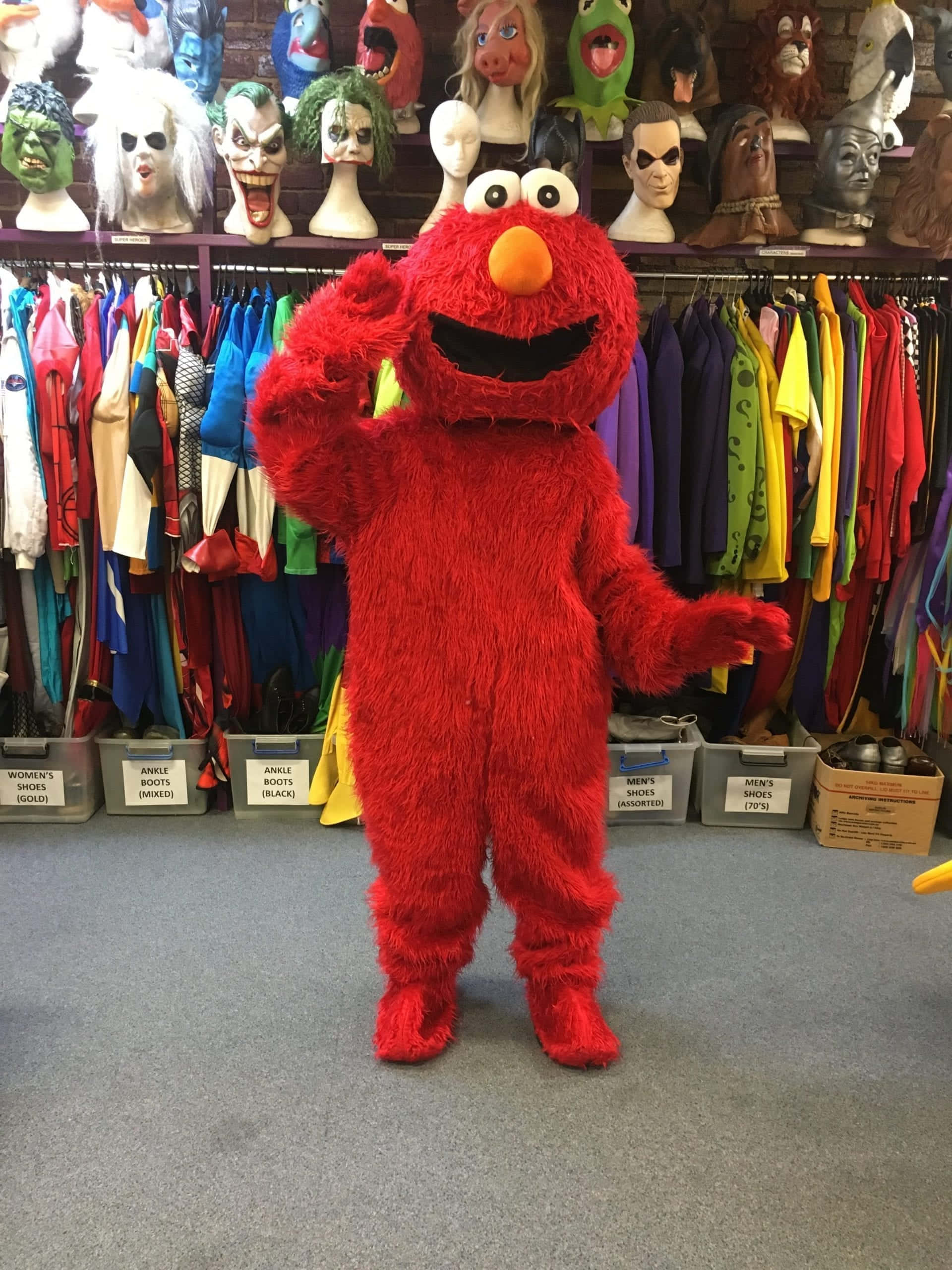 Sesame Street 桌布 壁紙 — 一個紅色毛茸茸的Elmo吉祥物在充滿色彩繽紛服裝和面具的服裝店裡愉快地揮手。