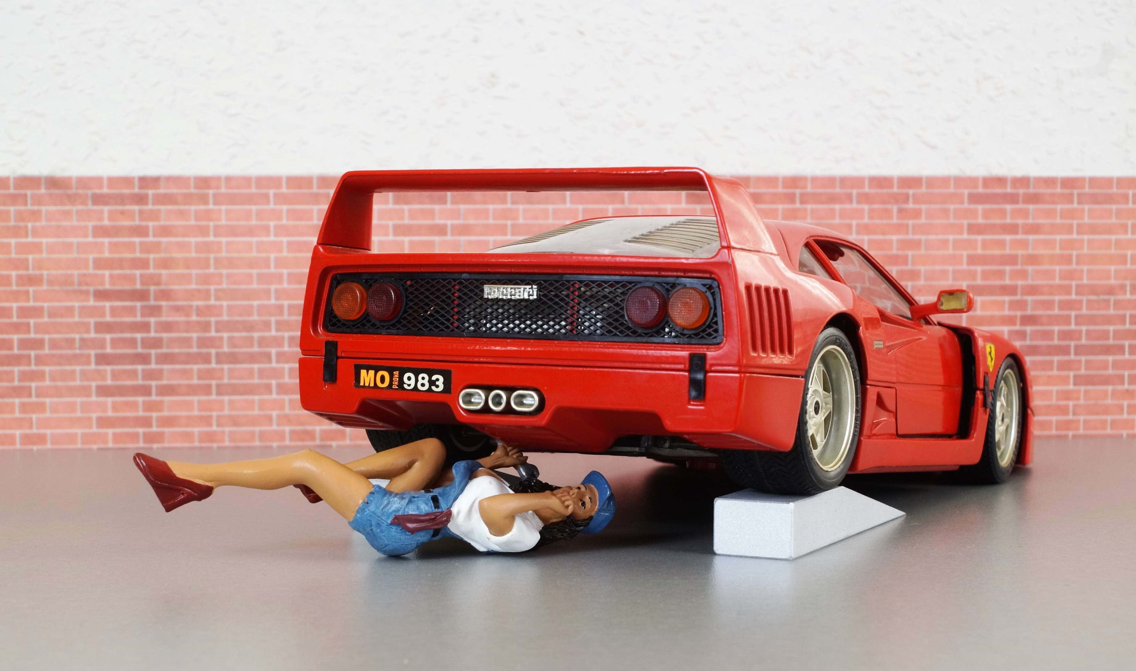 Vintage Ferrari 桌布 壁紙 — 一個紅色法拉利F40模型玩具，下面有一個公仔人物躺在灰色地板上，背景是磚牆。