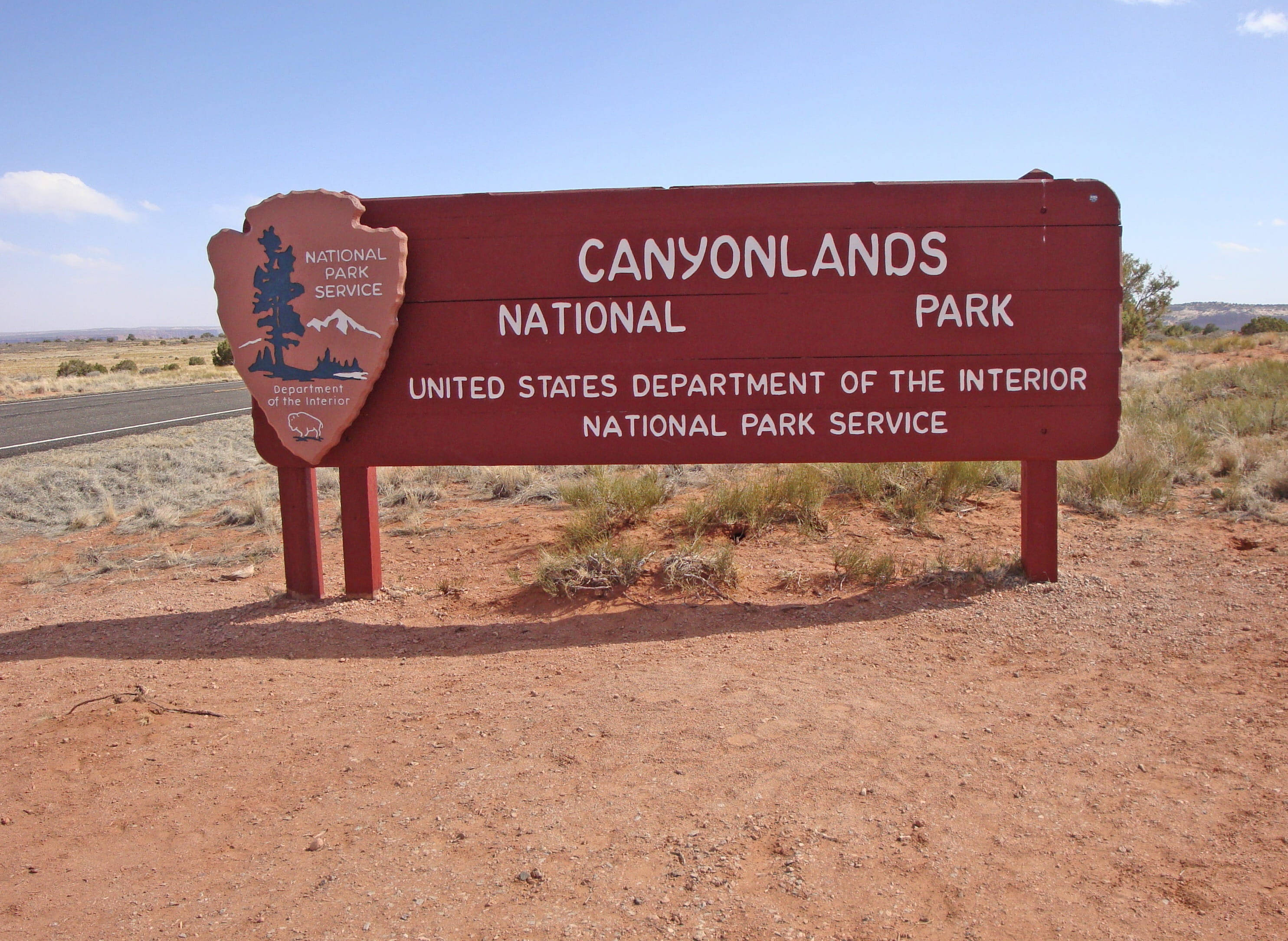 Canyonlands National Park 桌布 壁紙 — 一個紅色的峽谷地國家公園標誌，在乾燥的沙漠景觀中，在晴朗的藍天下，植被稀疏。