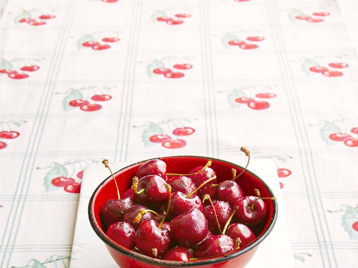 Red Cherries 桌布 壁紙 — 一個紅色碗裝滿了光澤的櫻桃，放在櫻桃圖案的桌布上，在明亮的日光下。