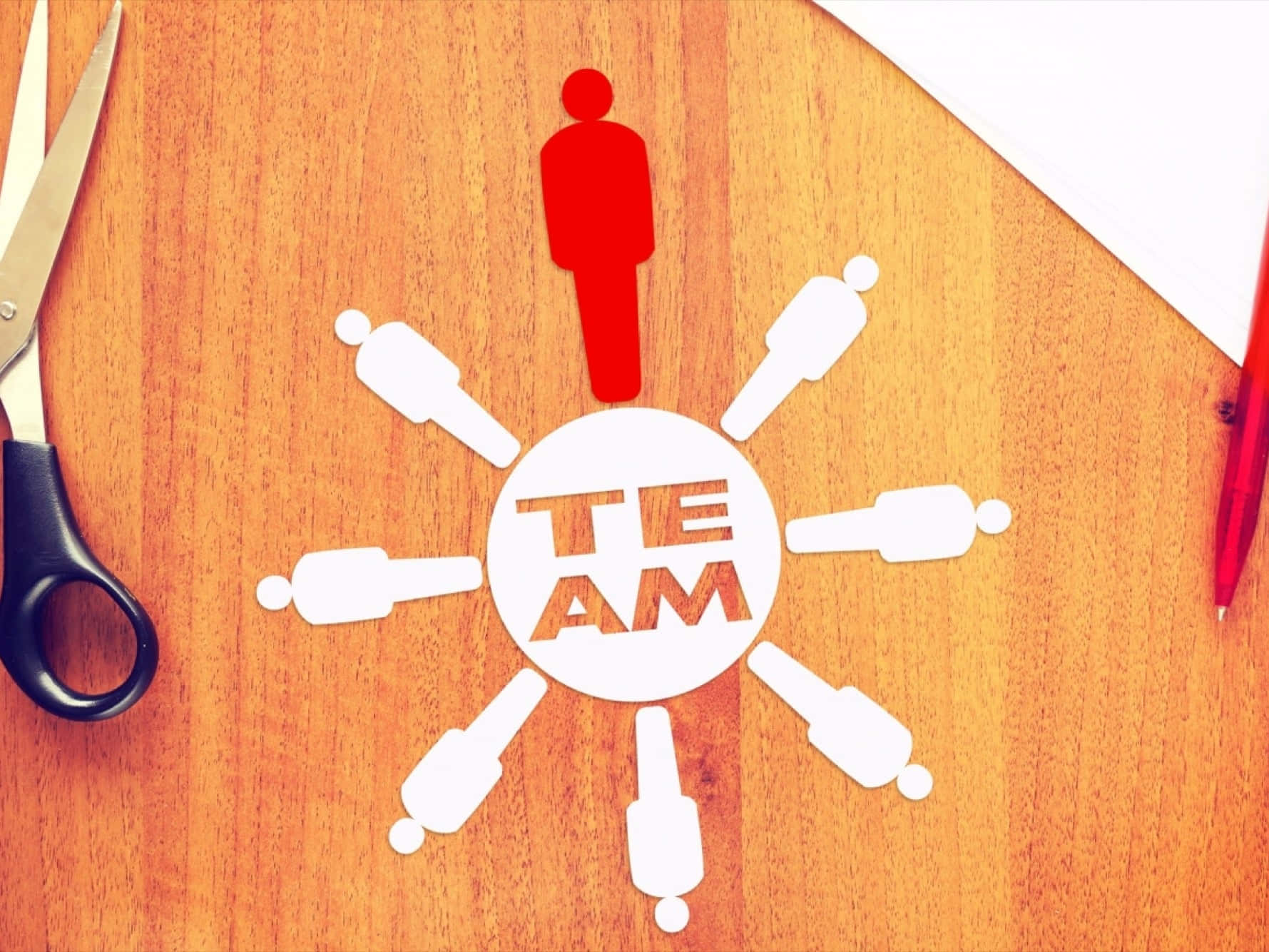 Leadership 桌布 壁紙 — 一個紅色紙剪出的人站在圓形「TEAM」標誌上，周圍有白色人物，木桌上附近有剪刀和筆。