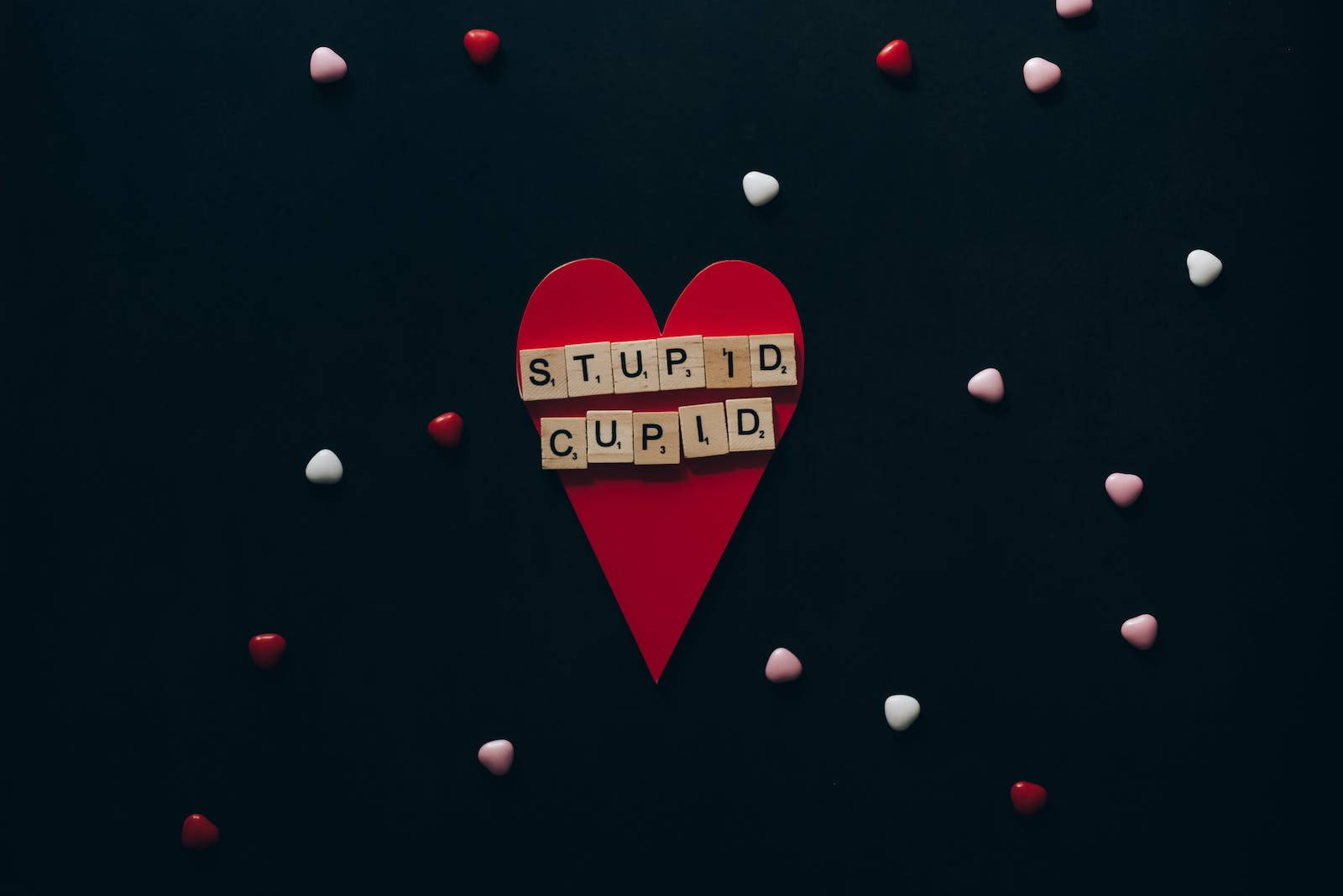 Stupid 桌布 壁紙 — 一個紅色紙心，上面有木製拼字板拼出「STUPID CUPID」，周圍散落著紅色和白色的心形，背景為深色。