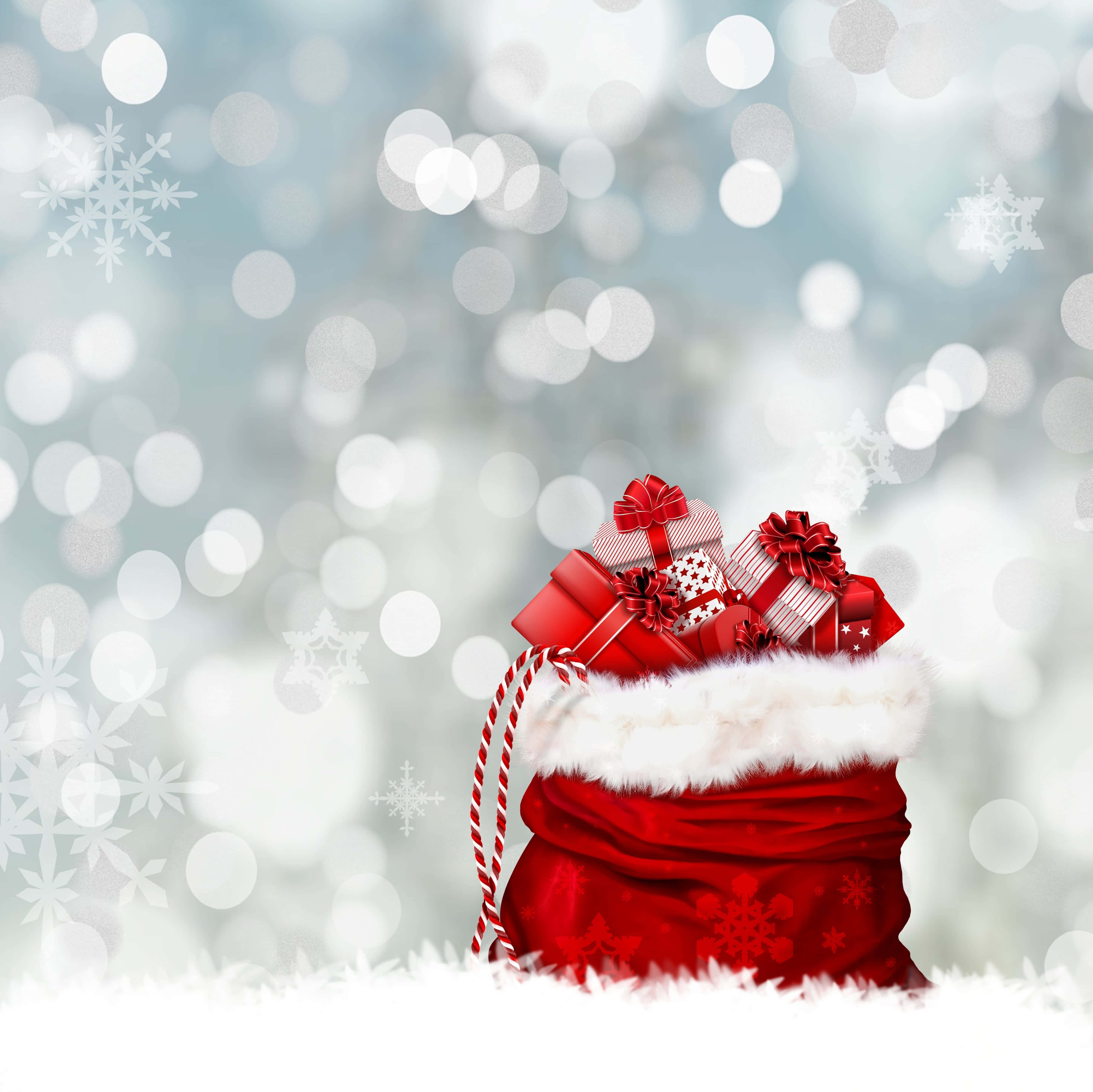 Gift 桌布 壁紙 — 一個紅色聖誕老人袋，裝滿了包裝好的禮物，放在雪地上，背景是散景的冬日景色和雪花。