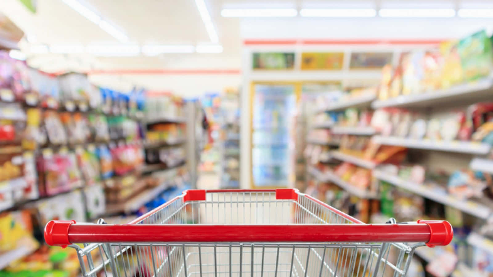 Shopping 桌布 壁紙 — 一個紅色購物車空置在光線明亮的雜貨店走道中，背景是模糊的彩色產品貨架。