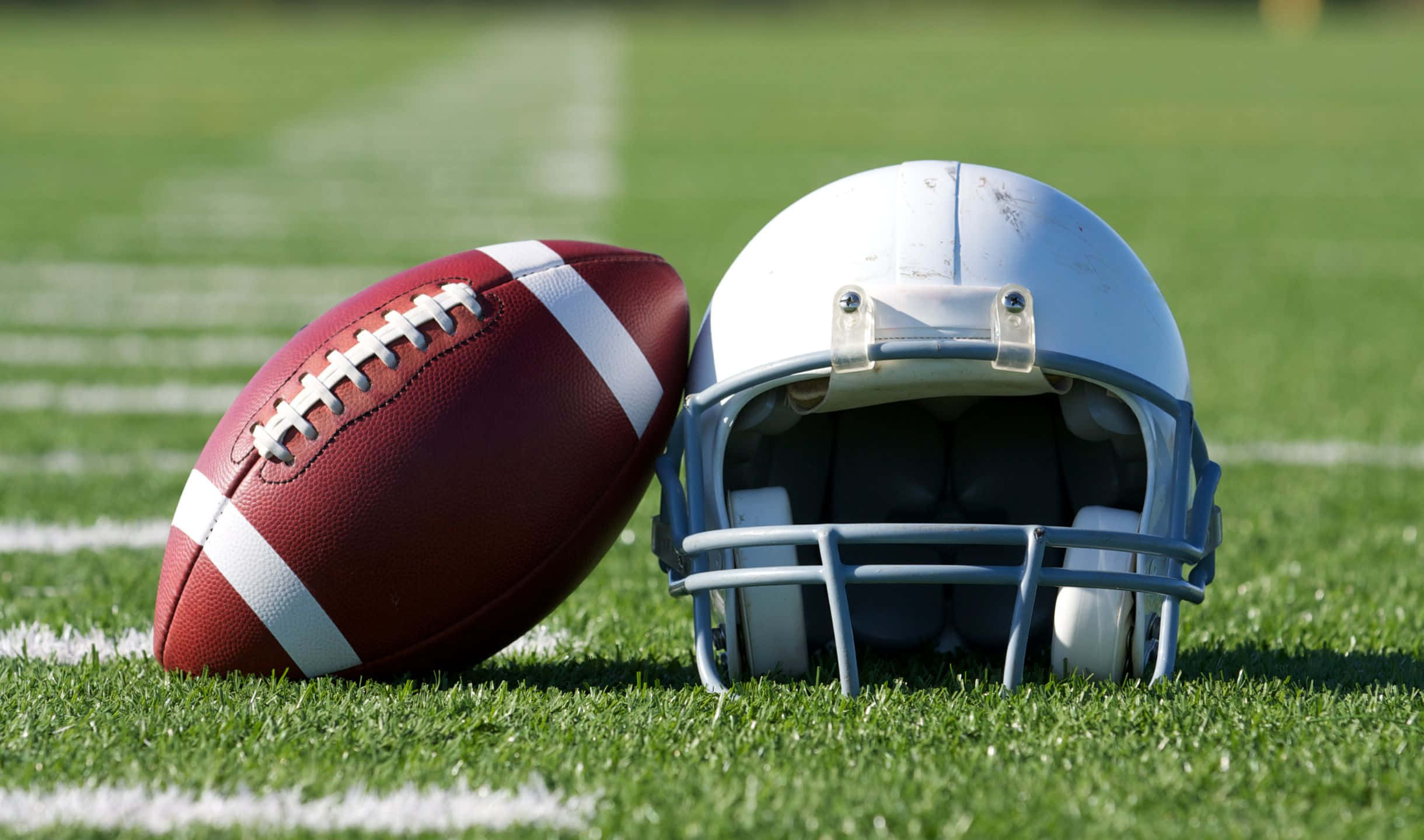2440x1440 Football 桌布 壁紙 — 一個紅色足球和白色頭盔放在鮮綠色草地上，草地有清晰的白線，在明亮的日光下。