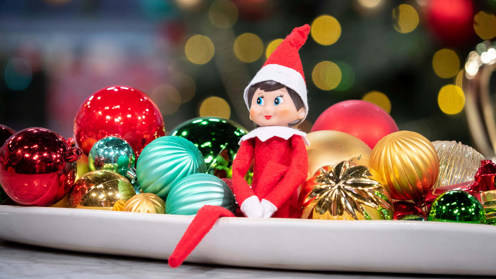 Christmas Elf 桌布 壁紙 — 一個紅色開心精靈公仔坐在白色托盤上，周圍是閃閃發光的多彩聖誕裝飾，背景有散景燈光。