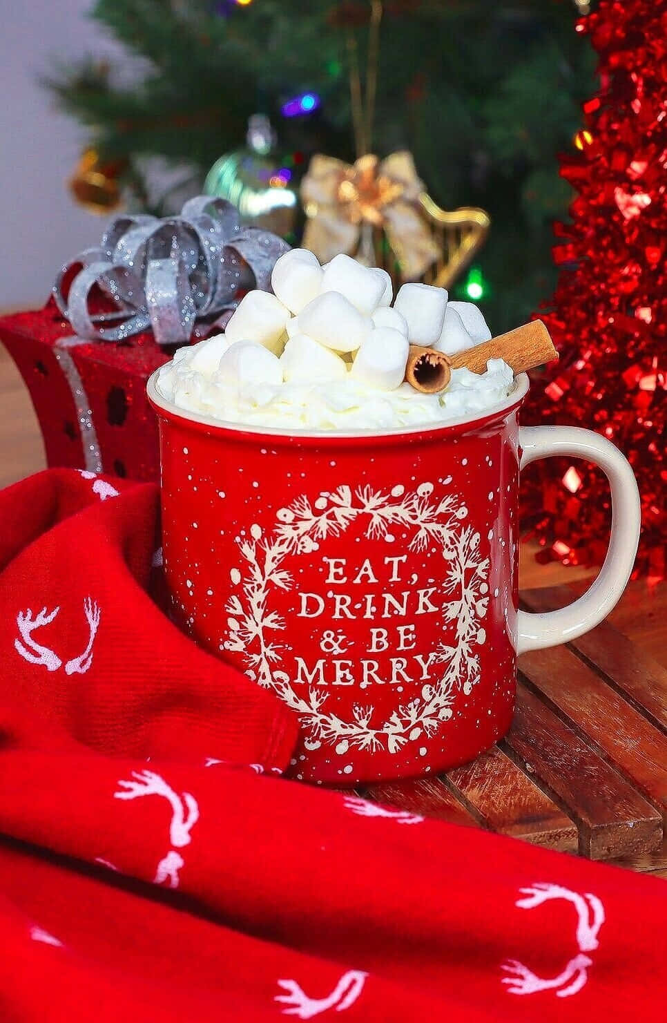 Hot Chocolate 桌布 壁紙 — 一個紅色馬克杯溢出鮮奶油和棉花糖，放在木桌上，旁邊有聖誕樹和禮物。