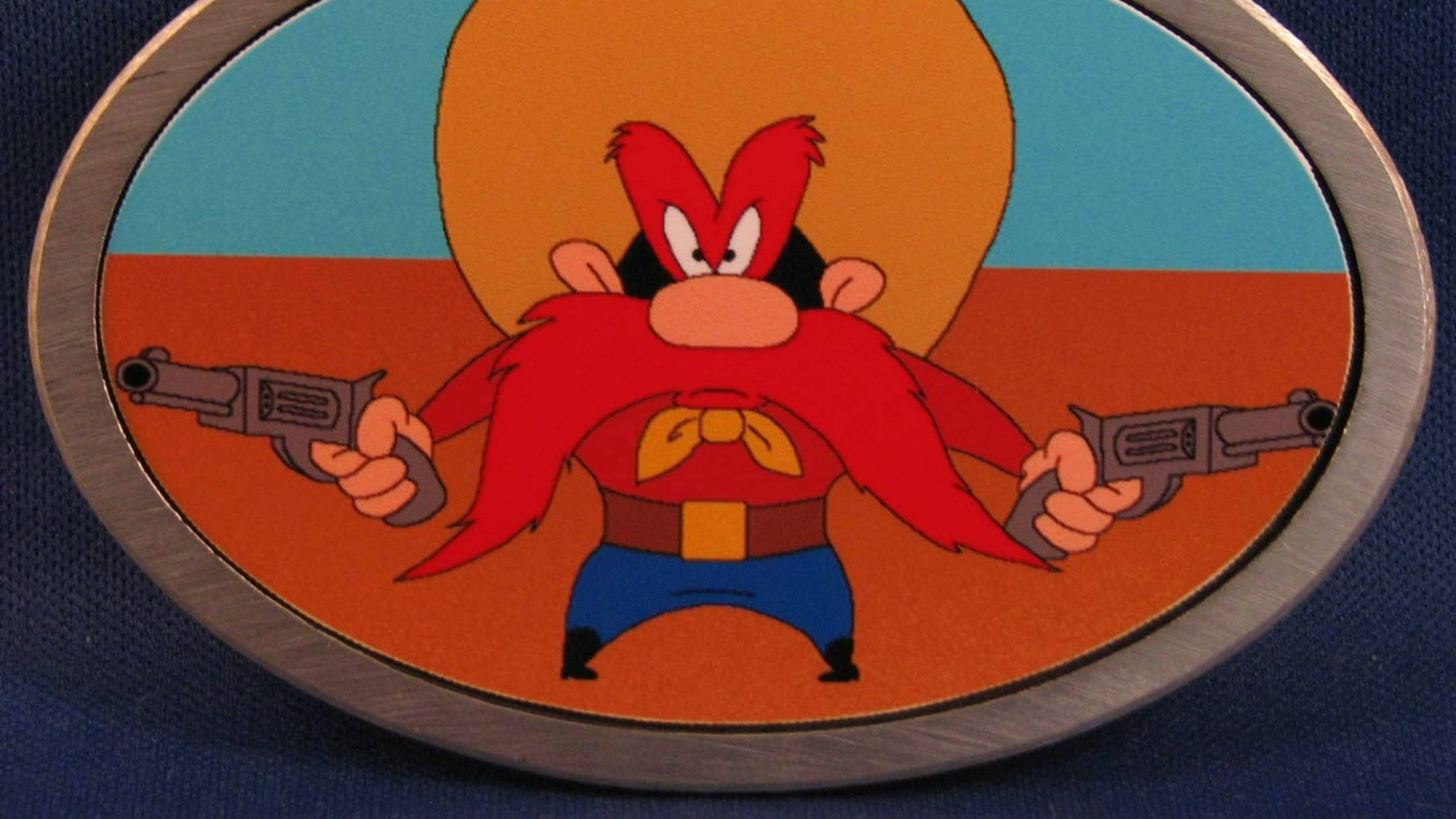 Yosemite Sam 桌布 壁紙 — 一個紅色鬍子和頭髮的卡通角色，在藍色布料背景上，手持兩把手槍放在銀色橢圓形扣環中。
