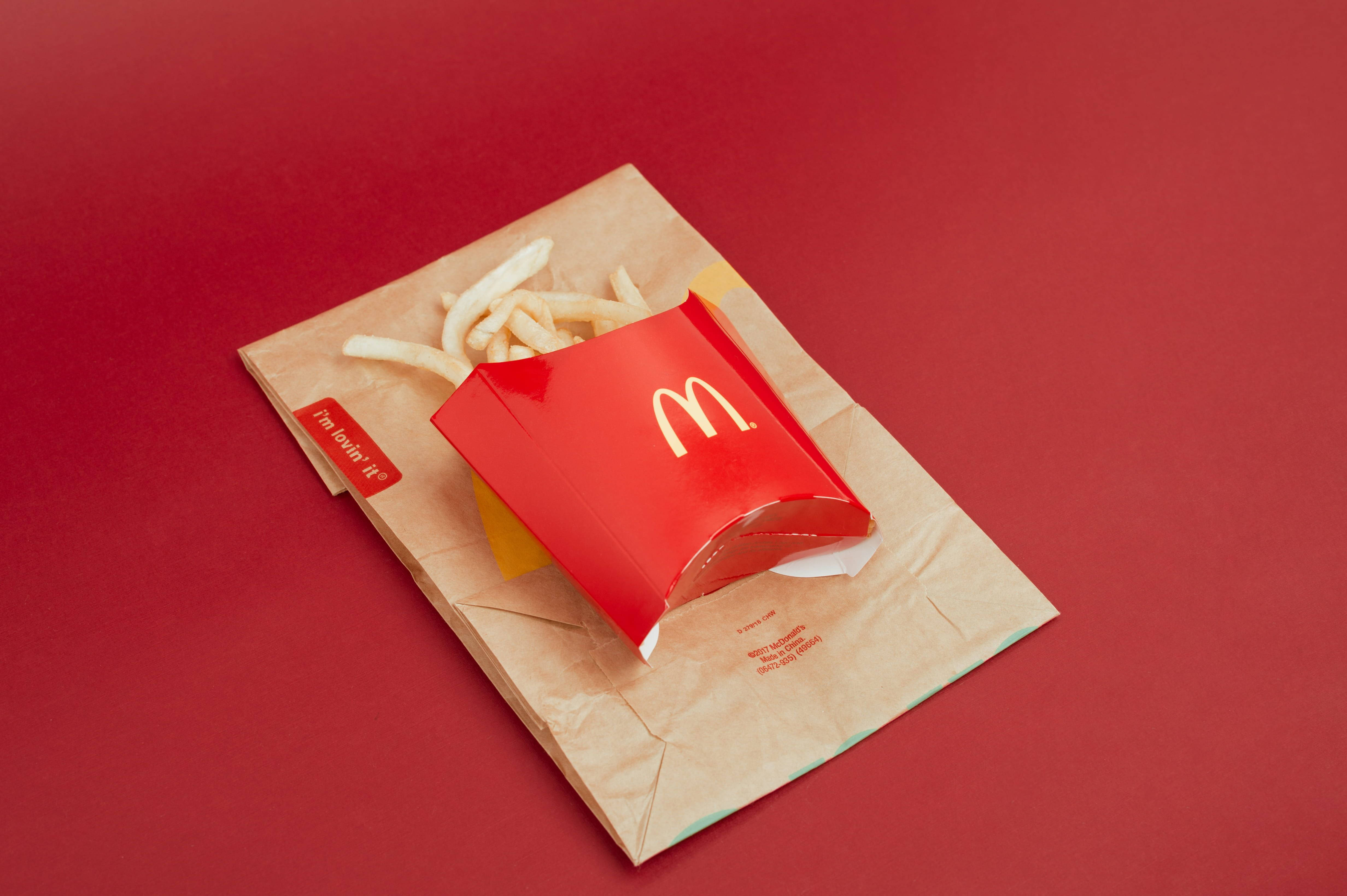 Mcdonalds 桌布 壁紙 — 一個紅色麥當勞薯條盒，裝滿金黃色薯條，放在一個棕色紙袋上，背景為純紅色。
