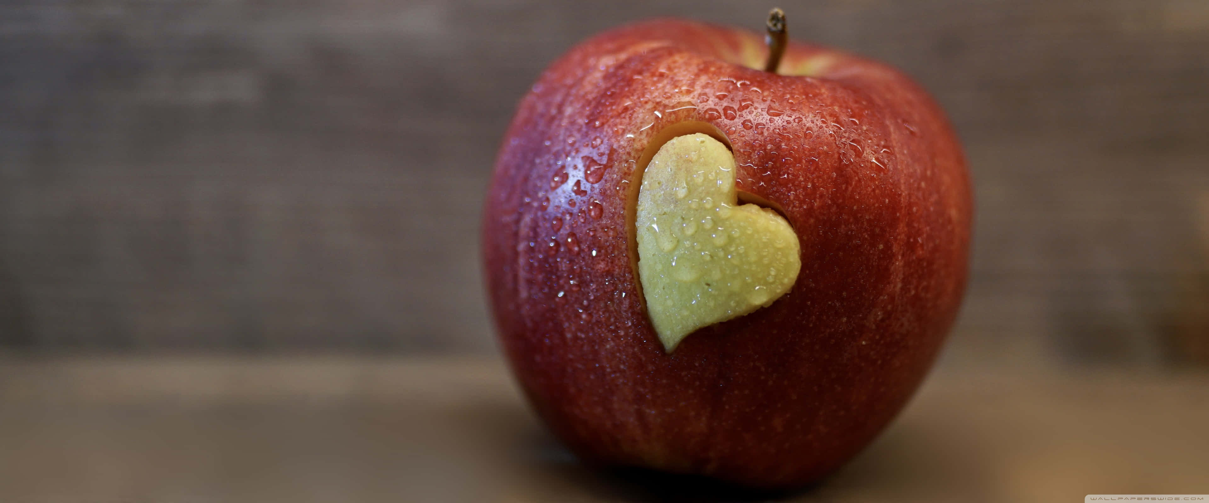 Apple 3840x1600 桌布 壁紙 — 一個紅蘋果的特寫，上面有一個心形的咬痕，露出淡黃色的果肉，背景是模糊的木頭。