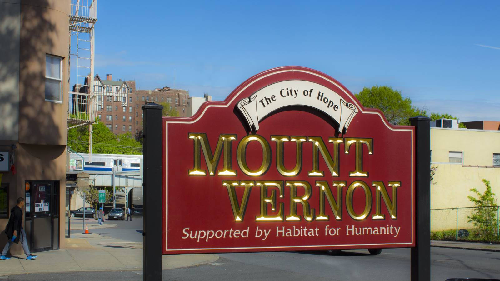 Blue Sky 桌布 壁紙 — 一個紅金色的 Mount Vernon 城市標誌在陽光明媚的街道上顯眼地豎立，背景有建築物和樹木。