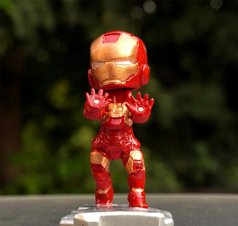 Iron Man Bobbleheads 桌布 壁紙 — 一個紅金色的Q版鋼鐵人人偶，站在透明底座上，雙手發出光芒，背景是模糊的綠色。