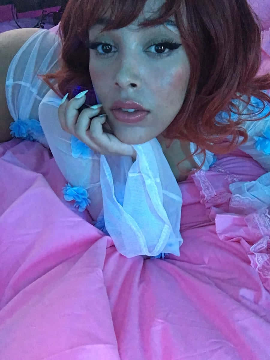 Doja Cat 桌布 壁紙 — 一個紅髮、妝容誇張的人躺在粉紅色床單上，自拍時將電話貼在耳邊。