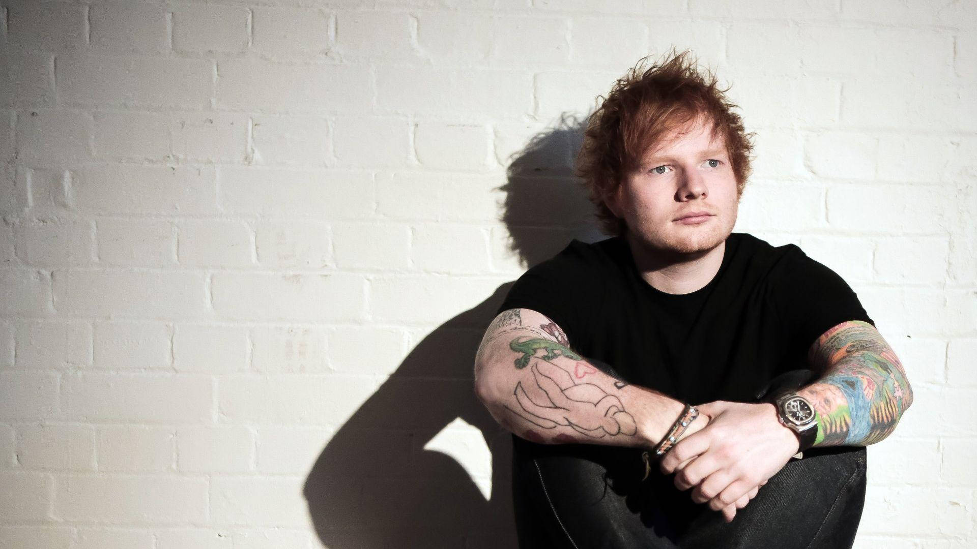 Ed Sheeran 桌布 壁紙 — 一個紅髮、身上有彩色刺青的男人靠著白色磚牆，在強烈的陽光下投下清晰的影子。