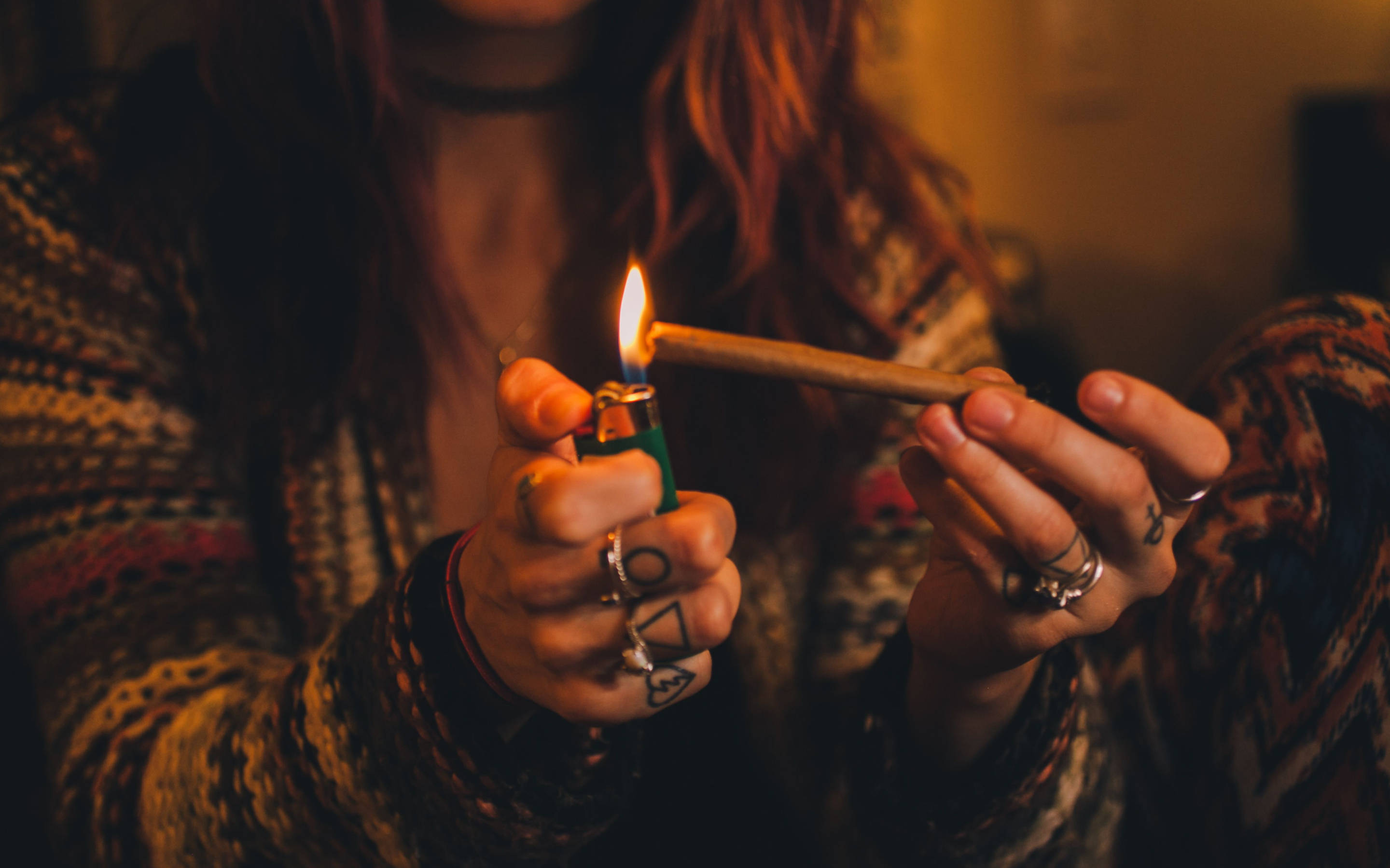 吸大麻 桌布 壁紙 — 一個紅髮且有刺青的人，在昏暗溫暖的房間裡用綠色打火機點燃大麻菸。