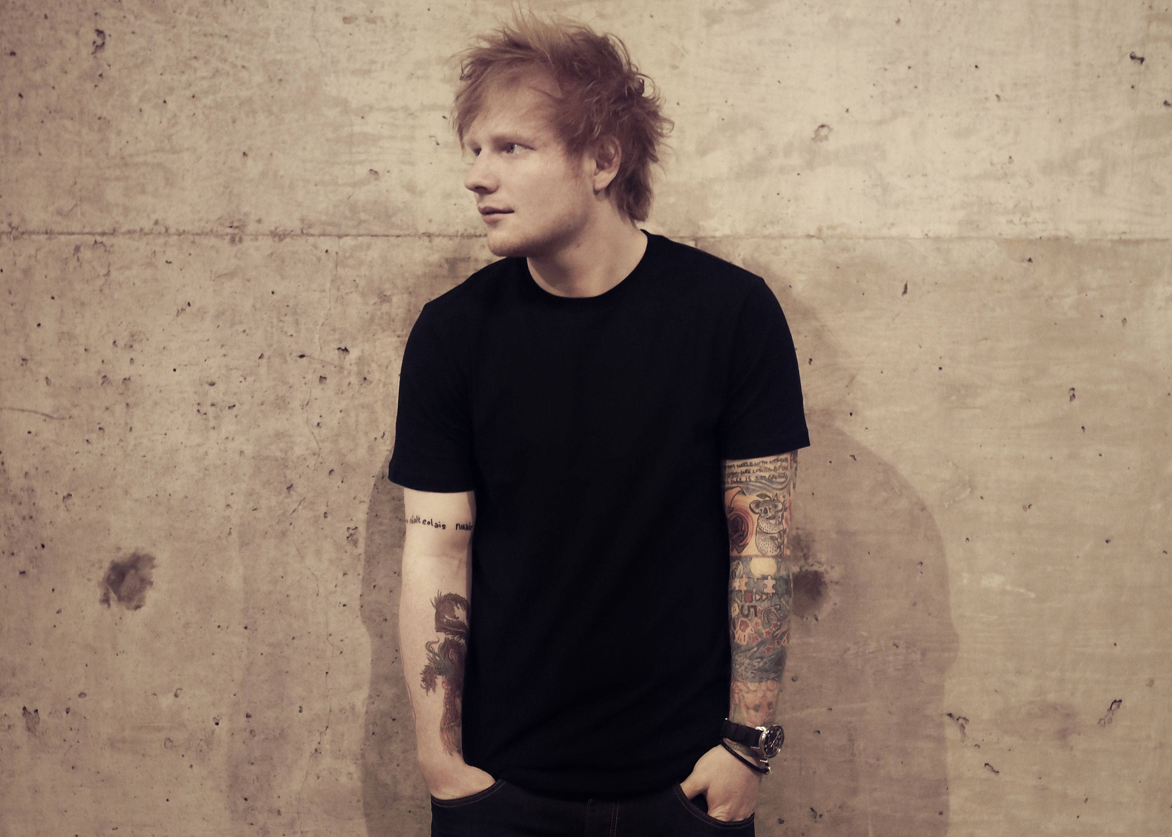 Ed Sheeran 桌布 壁紙 — 一個紅髮刺青男子穿著黑色T恤靠著紋理混凝土牆。
