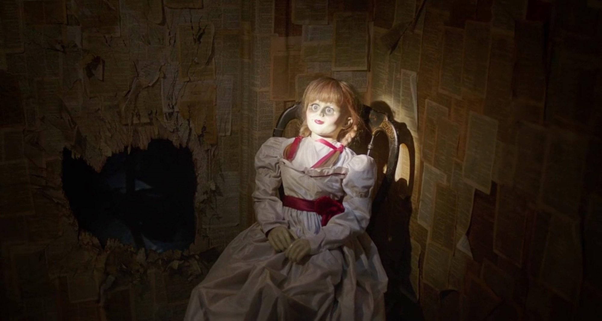 Annabelle 桌布 壁紙 — 一個紅髮玻璃眼的恐怖娃娃坐在椅子上，周圍是撕破的報紙牆，房間光線昏暗。