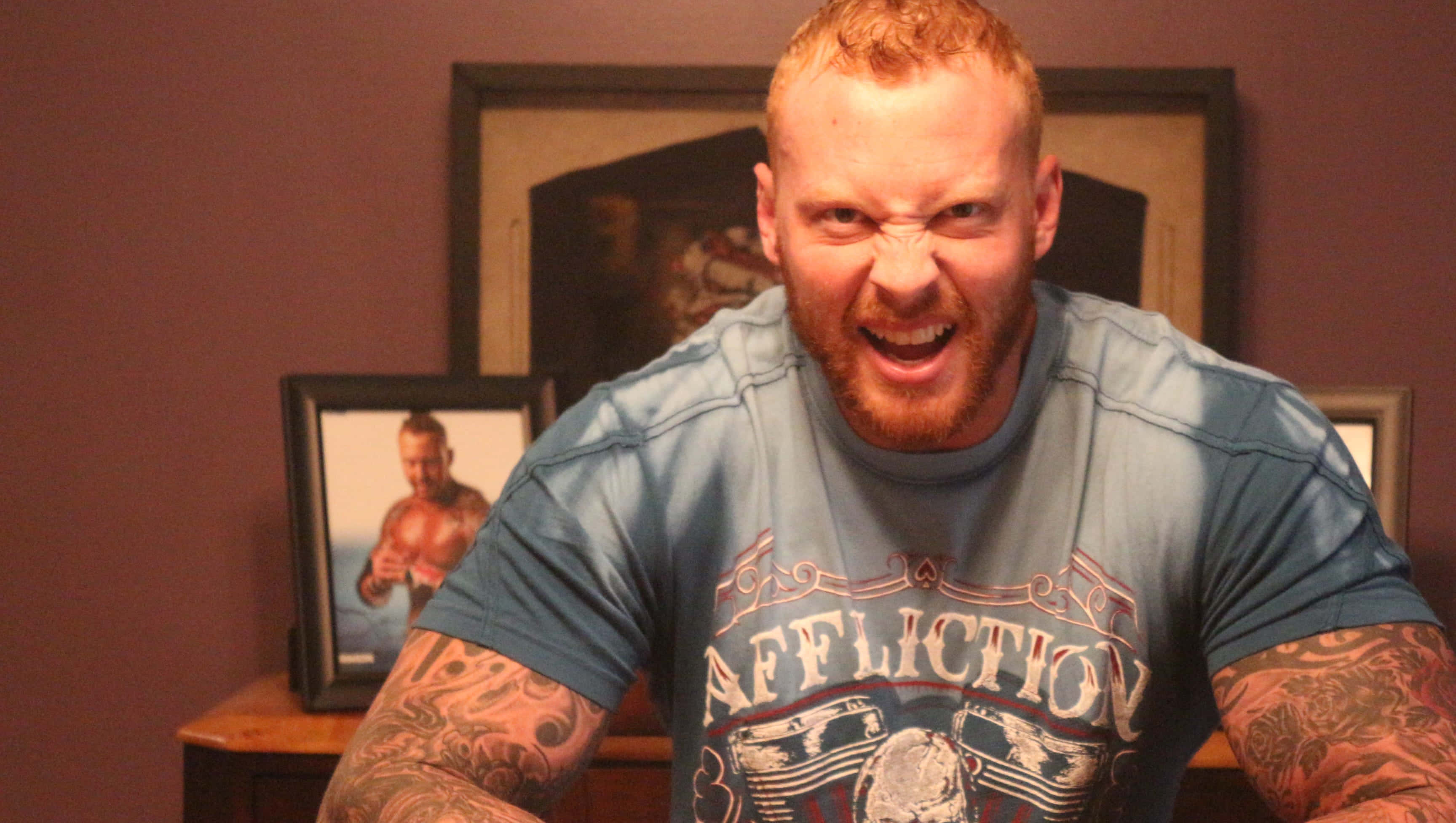 摔角 桌布 壁紙 — 一個紅髮紋身的肌肉男在室內穿著藍色 'Affliction' T恤，表情猙獰。