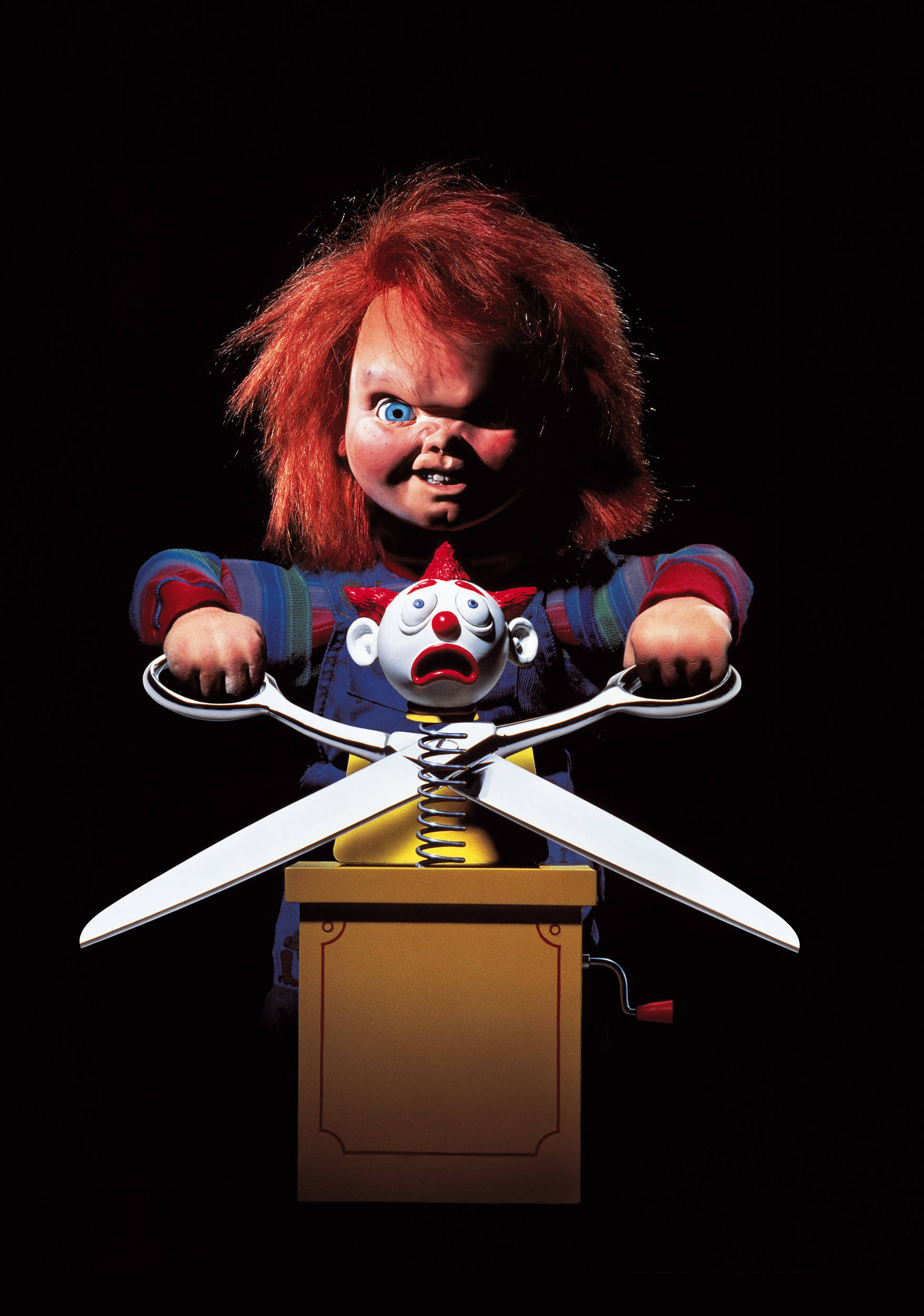 Chucky 桌布 壁紙 — 一個紅髮藍眼的恐怖娃娃在黑色背景下的木箱中，手持巨型剪刀對準小丑頭部。