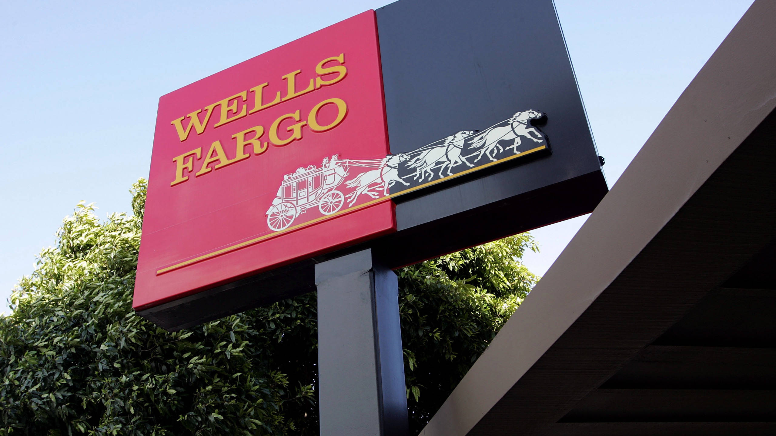 樹木 桌布 壁紙 — 一個紅黑相間的 Wells Fargo 標誌，帶有金色馬車標誌，在藍天白雲和下方綠樹的襯托下格外醒目。