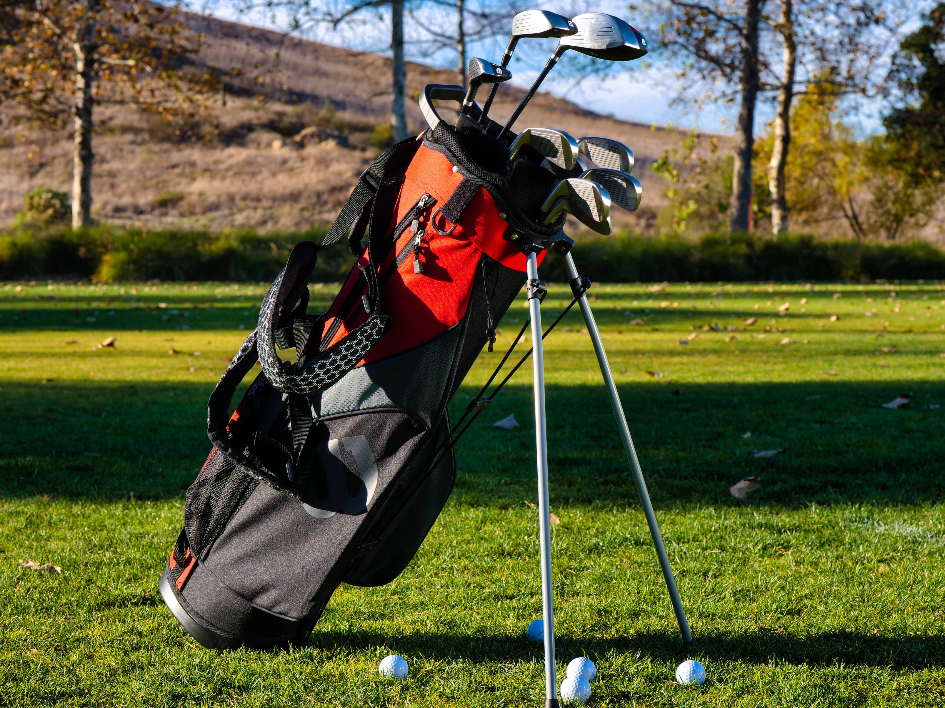 4K Golf 桌布 壁紙 — 一個紅黑相間的高爾夫球袋直立於陽光普照的綠色球道上，球桿露出，附近散落著高爾夫球。