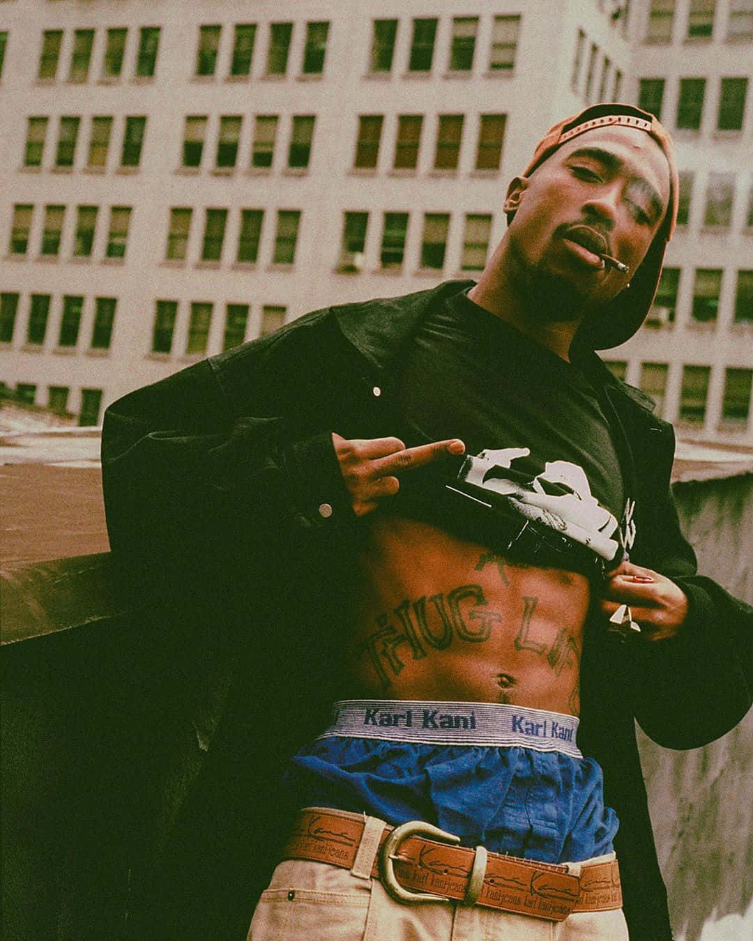 tupac 桌布 壁紙 — 一個紋身上身、口中叼著雪茄的男人，自信地在城市建築背景下擺姿勢。