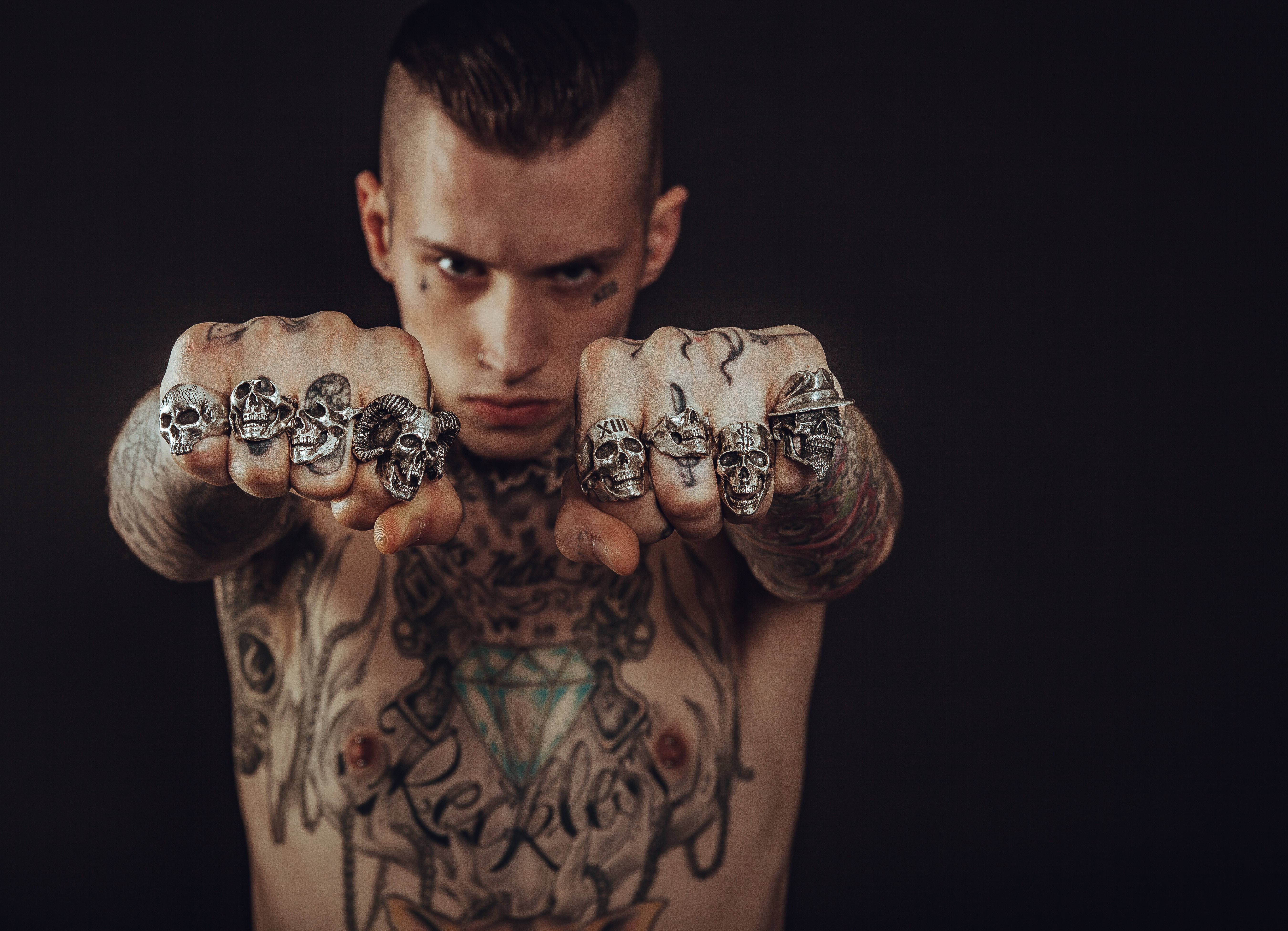 Tattoo 桌布 壁紙 — 一個紋身男子，眼神銳利，戴著骷髏戒指，在黑暗背景前直視前方。