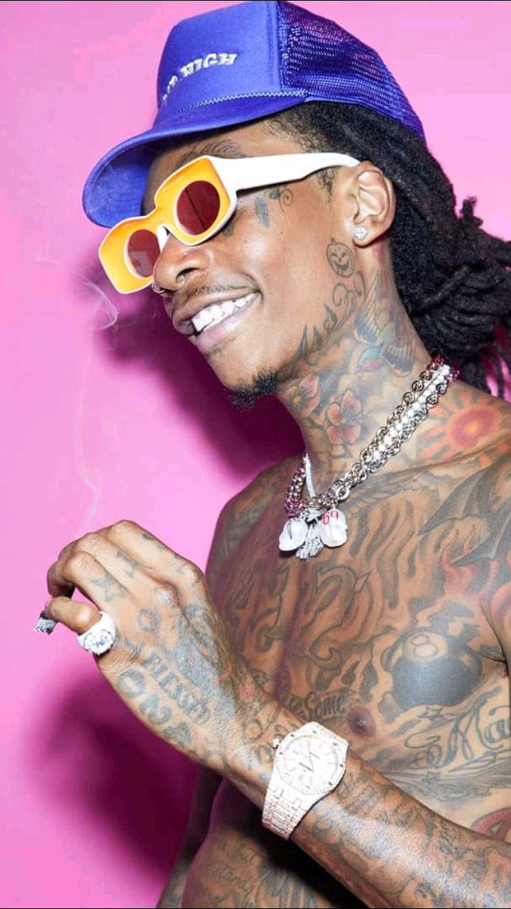 wiz khalifa 桌布 壁紙 — 一個紋身男子戴著藍色帽子和黃色太陽眼鏡，在充滿活力的粉紅色背景前吸煙。