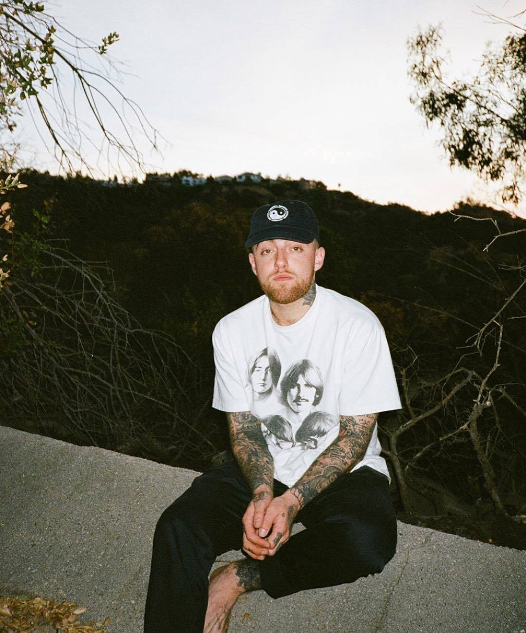 Mac Miller 桌布 壁紙 — 一個紋身男子戴著黑色帽子，身穿白色Beatles T恤，在黃昏時坐在混凝土邊緣，無葉的樹枝以丘陵背景襯托著他。