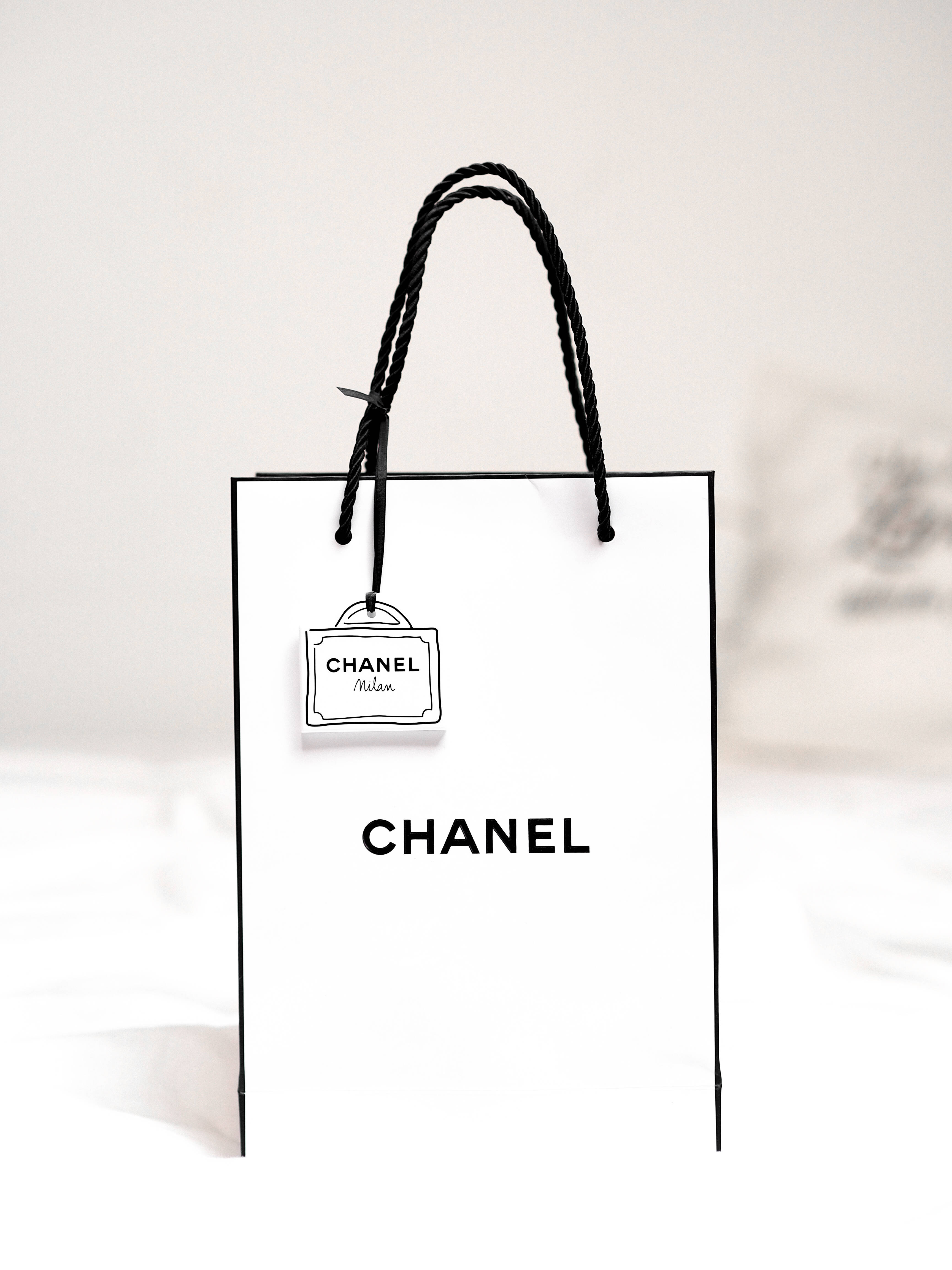 Gucci 桌布 壁紙 — 一個純白色的Chanel購物袋，帶有黑色手柄和標誌，置中於柔和的模糊背景上。