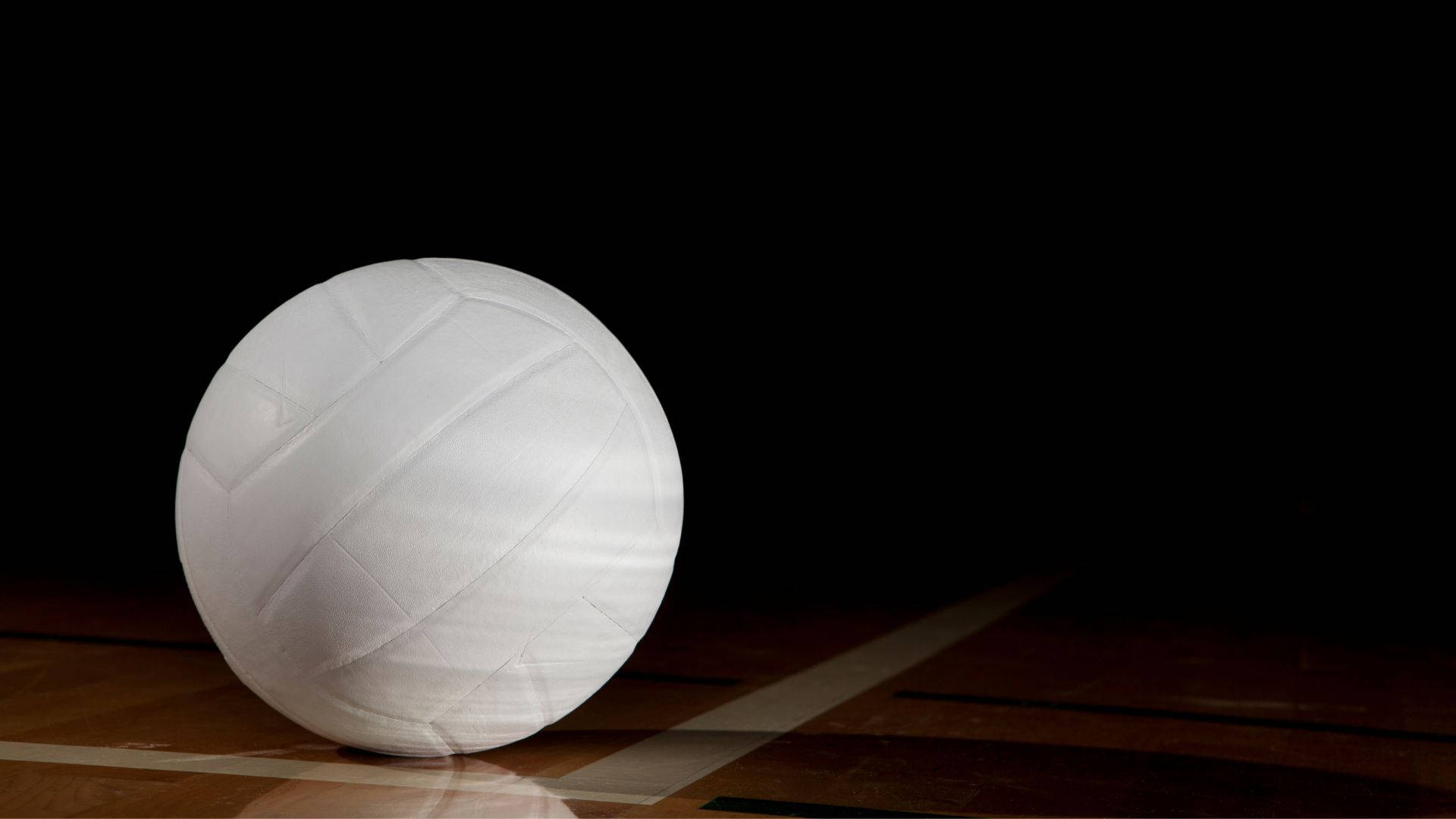 Volleyball Aesthetic 桌布 壁紙 — 一個純白色的排球放在拋光木地板上，在黑色背景下受到戲劇性的聚光燈照射。