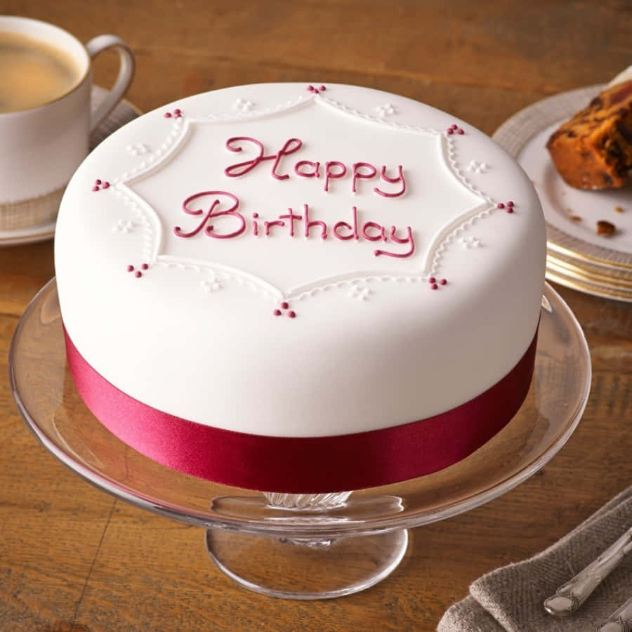 Happy Birthday Cake 桌布 壁紙 — 一個純白色的生日蛋糕，上面有紅色絲帶和文字糖霜，放在玻璃架上，旁邊有一杯咖啡和糕點。