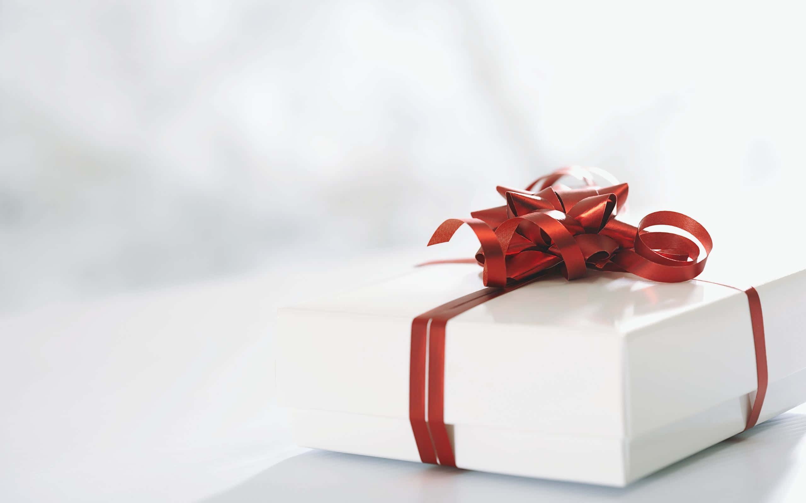Gift 桌布 壁紙 — 一個純白色的禮物盒，飾有光澤的紅色絲帶蝴蝶結，放在明亮的白色表面上。