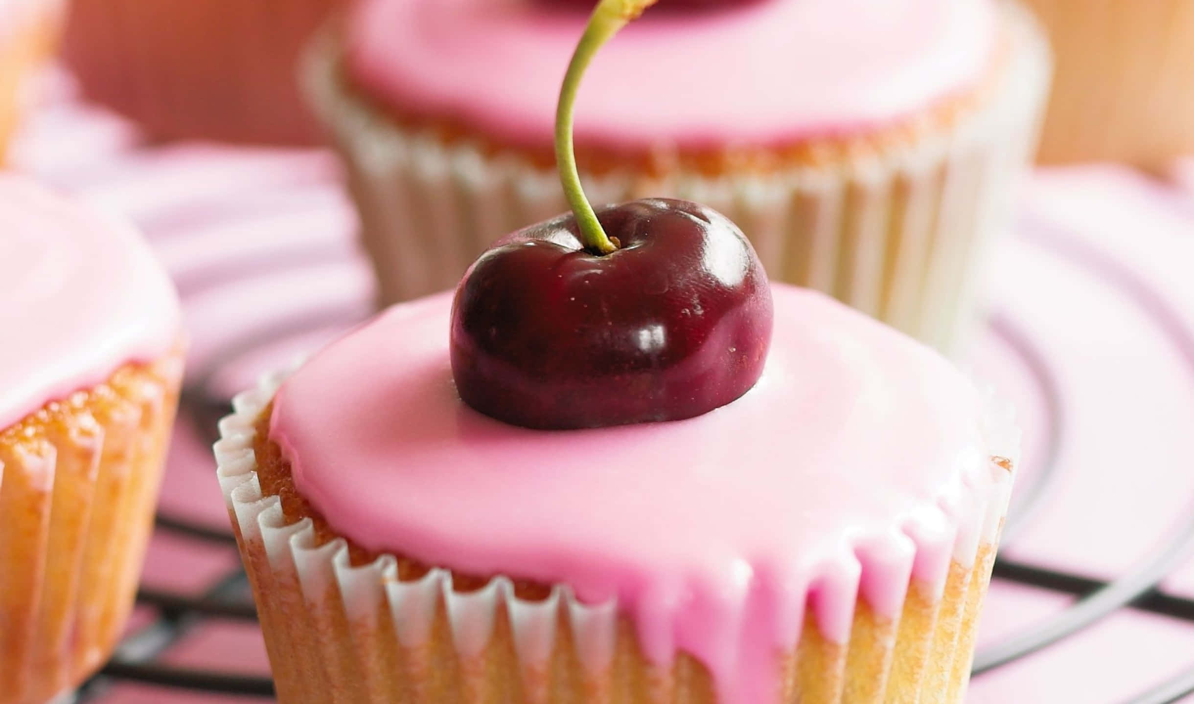 2440x1440 Desserts 桌布 壁紙 — 一個紙杯蛋糕的特寫，上面有粉紅色糖霜和一個光亮的櫻桃，放在金屬架上。