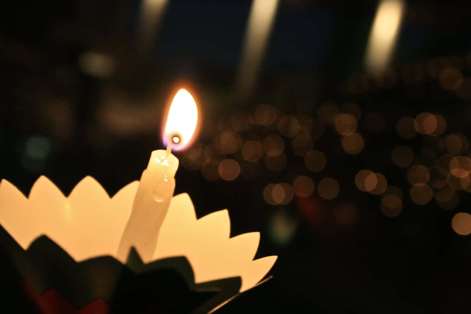 Candle Light 桌布 壁紙 — 一個紙製燭台上的單支點燃蠟燭，在帶有散景燈光的黑暗背景中散發出溫暖的光芒。
