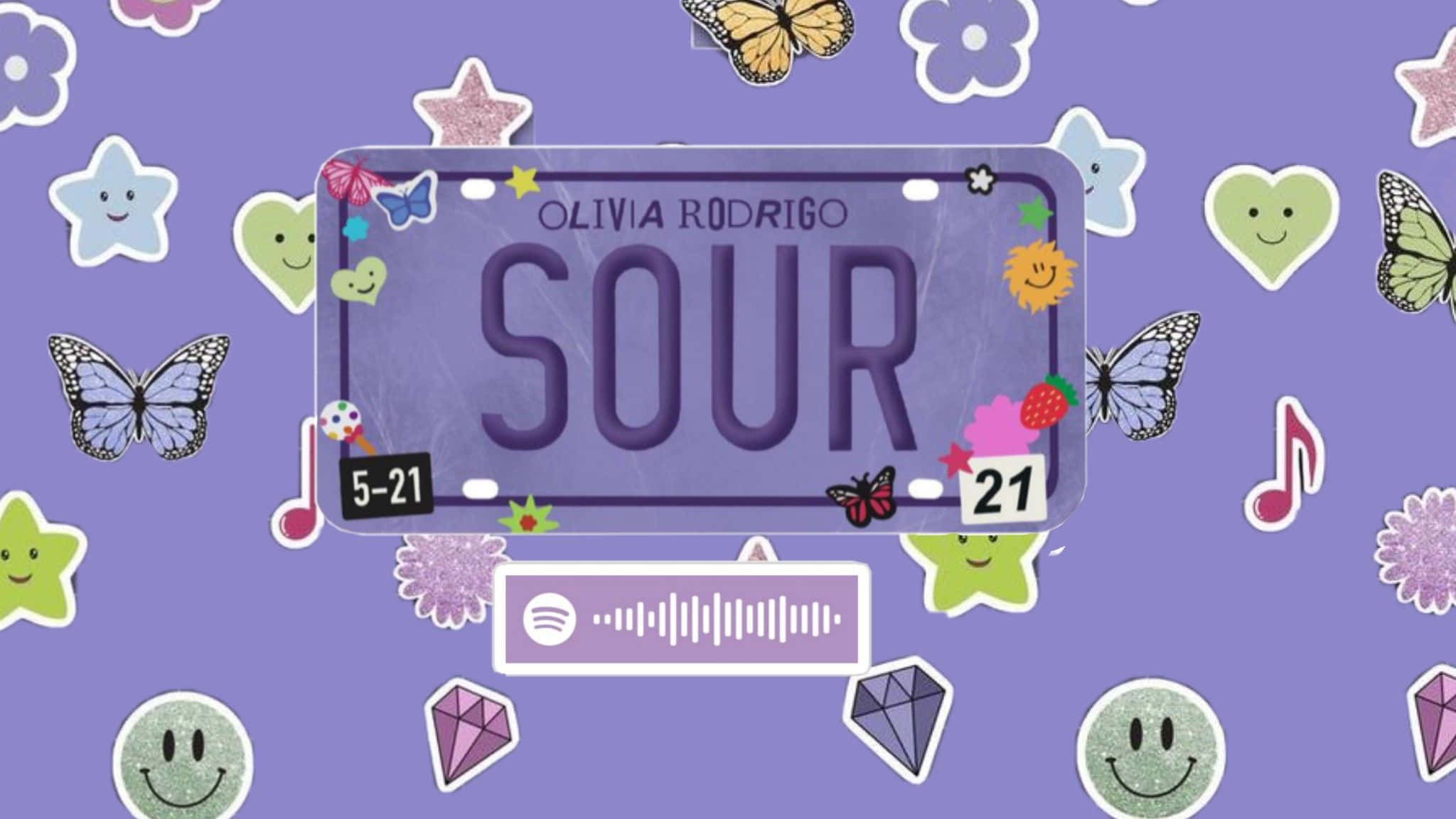 奧莉維亞·羅德里戈 桌布 壁紙 — 一個紫色車牌，上面寫著 Olivia Rodrigo 的「SOUR」，周圍有色彩繽紛的蝴蝶、愛心和音符貼紙，背景閃閃發光。