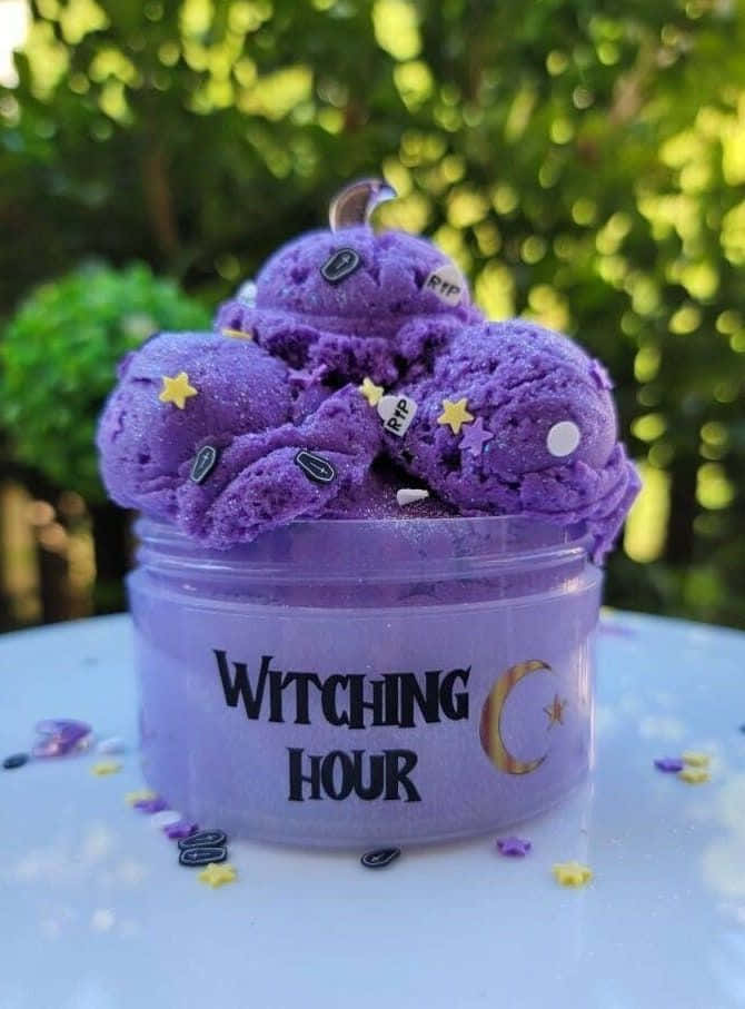 slime 桌布 壁紙 — 一個紫色閃閃發光的史萊姆，標有Witching Hour，上面有月亮和星星裝飾，放在白色表面上，背景是綠色模糊的花園。