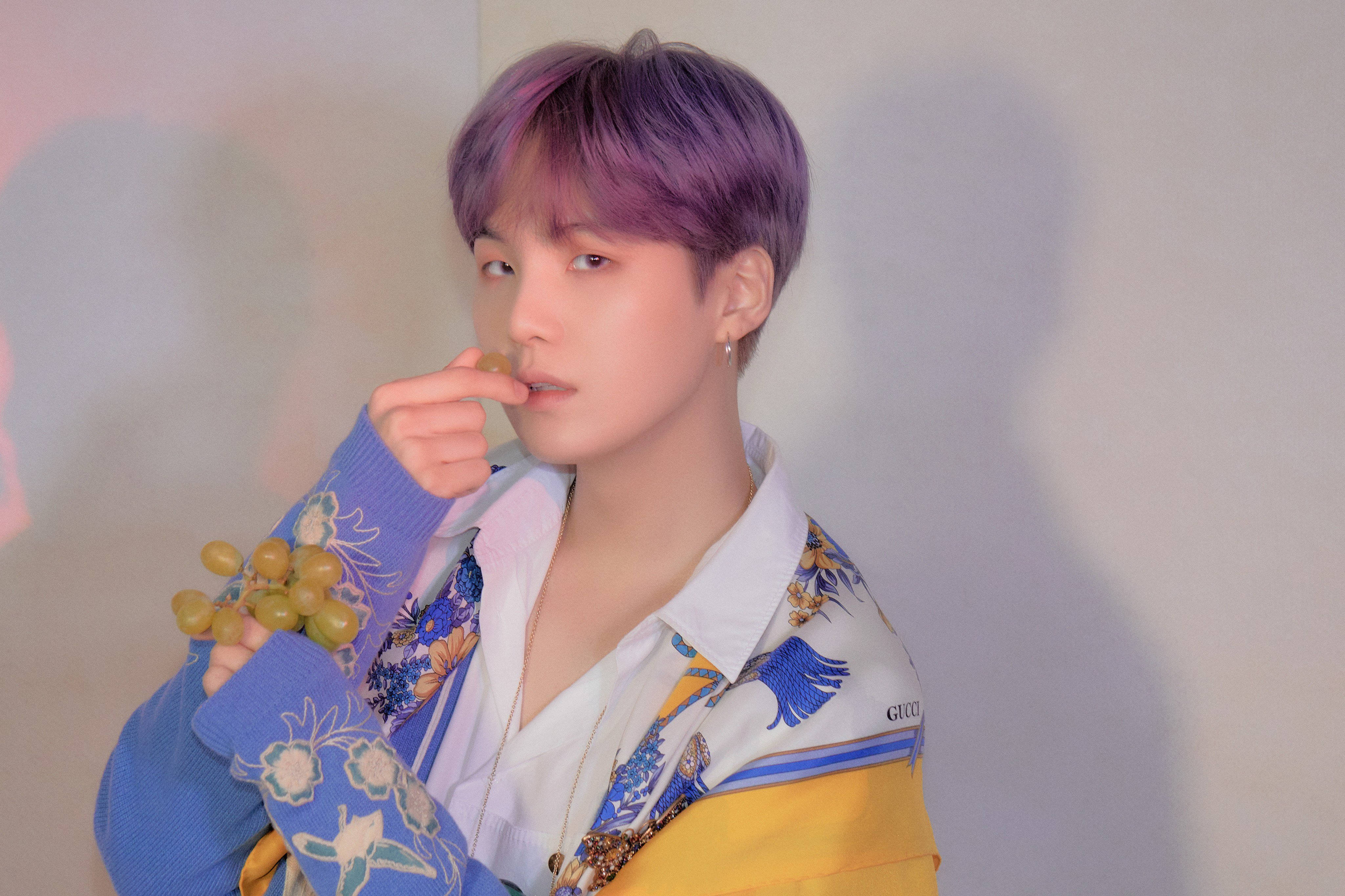 Suga Bts 桌布 壁紙 — 一個紫色頭髮的人拿著綠色葡萄，穿著彩色圖案外套，背景是素色牆壁。