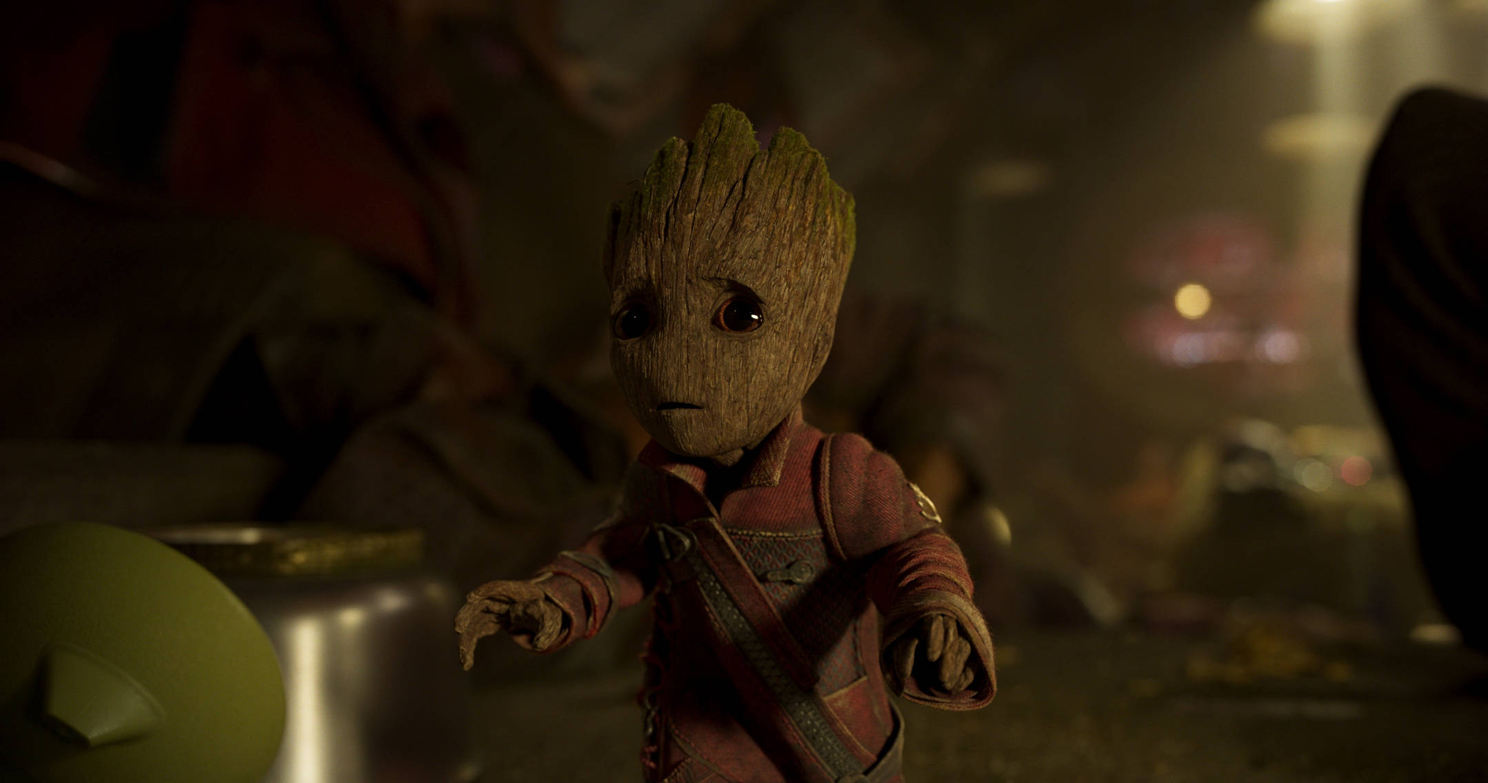 Baby Groot 桌布 壁紙 — 一個細小、像樹一樣的生物，有大眼睛，穿著紅色外套，站在一個光線昏暗、雜亂的房間裡，背景有溫暖發光的燈光。